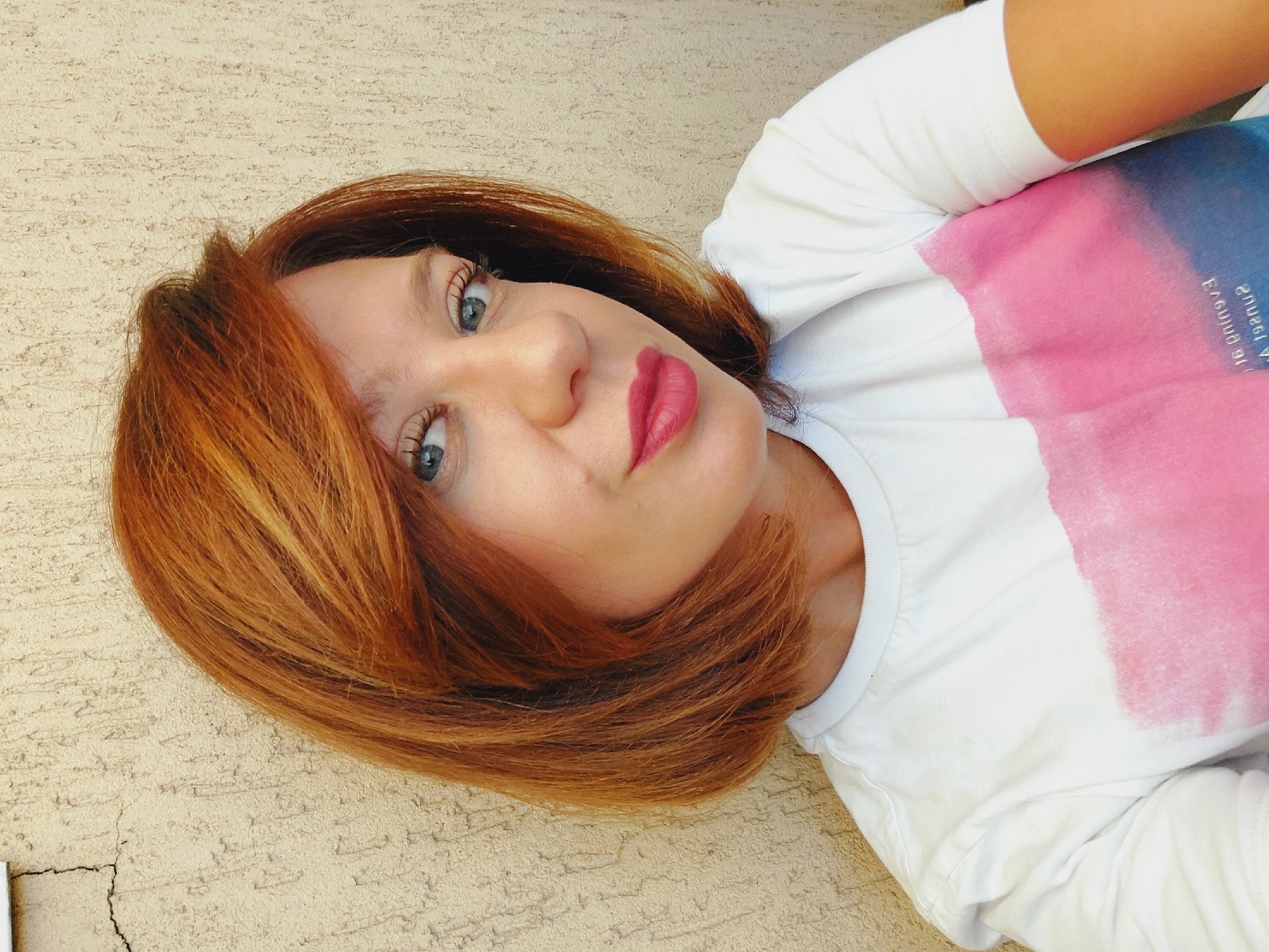 A medium close-up selfie shows a woman with auburn hair styled in a bob, wearing a white t-shirt with a pink and blue abstract design. She has blue eyes and is wearing pink lipstick. The background is a textured, light-colored wall. The image appears to be taken outdoors, with natural light.