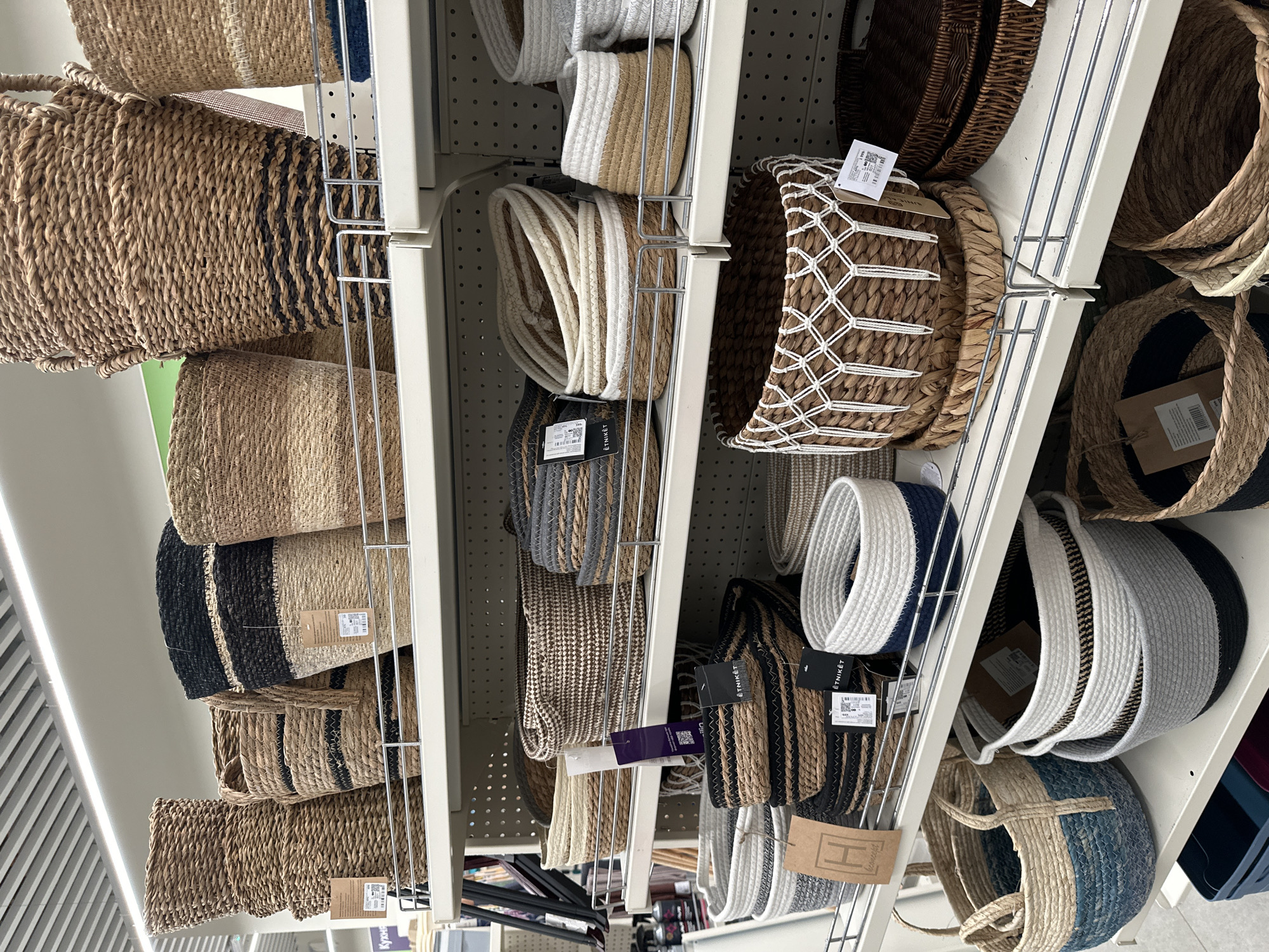 The image displays a retail setting, likely a home goods or decor store, with shelves stocked with various woven baskets. The shelves are metal and extend across the frame, holding multiple rows of baskets. The baskets vary in size, shape, material (primarily woven natural fibers like seagrass or jute), and color. Some baskets are plain, while others have decorative patterns, stripes, or contrasting colored bands. Several baskets have price tags or product information labels attached. In the background, the store's ceiling with fluorescent lighting fixtures and a perforated wall panel are visible. The overall impression is one of an organized retail display. There are no people visible in the image. The location is stated to be Samara, Russia, which is a city in Russia.