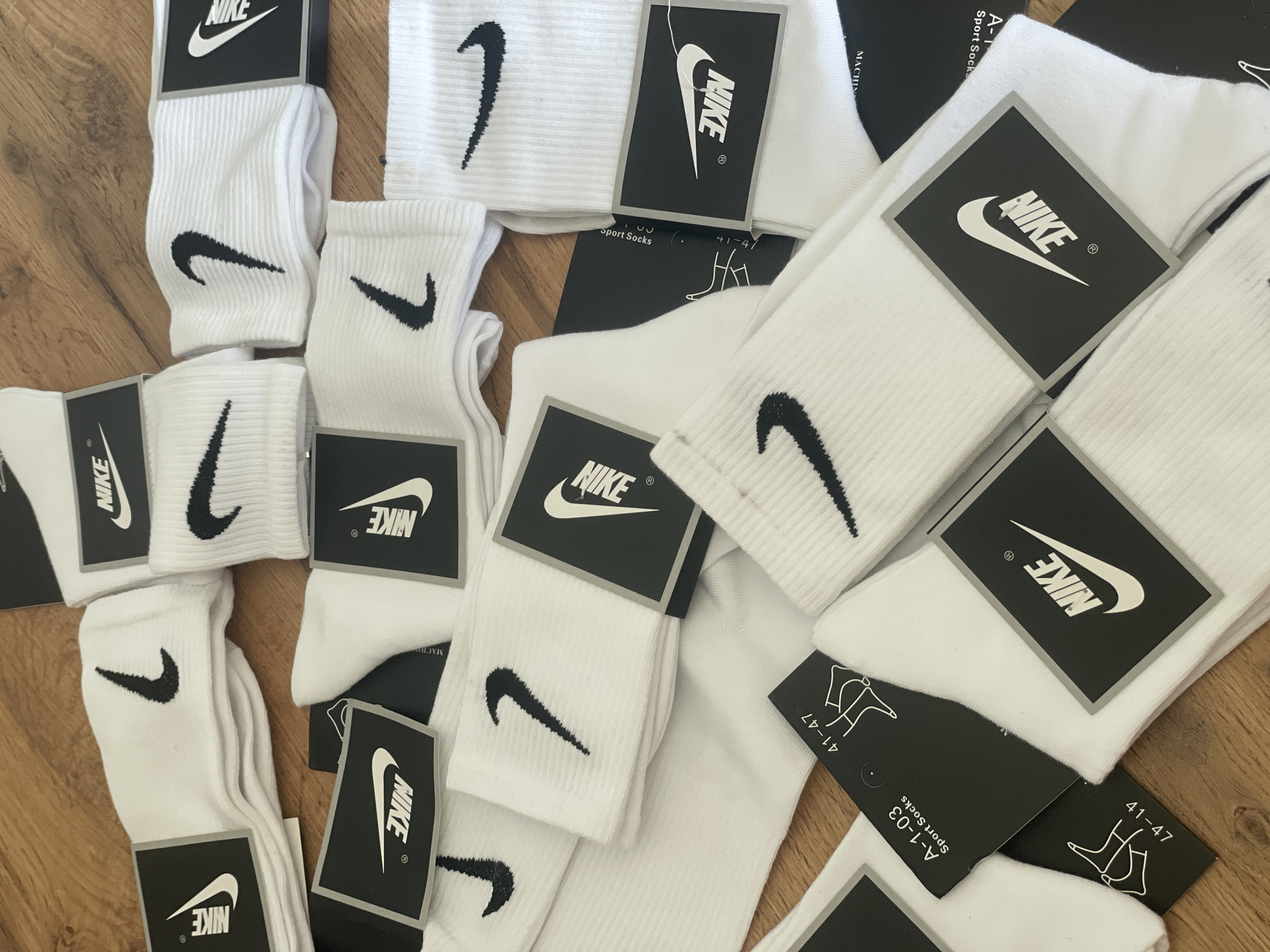 The image shows a pile of white Nike socks with black Nike logos, laid out on a wooden surface. Some of the socks are still in their original packaging with black tags displaying the Nike logo and size information, such as "41-47". The socks are neatly folded and stacked, with some appearing slightly creased. The lighting suggests an indoor environment. There are no people or specific landmarks visible to indicate a location in Krasnyi Krym, Russia.