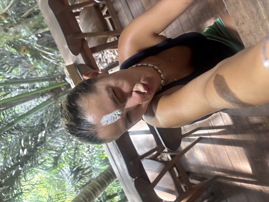 A single adult female is captured in a close-up selfie, positioned in an open-air, tropical environment consistent with the city of Punggul, Indonesia. She has damp, light-colored hair and a prominent vertical stripe of white, pasty substance, possibly clay, on her forehead, containing a small, lumpy gray object. Her right eye is closed in a wink, and her lips are pursed. She is wearing a dark tank top, a shell necklace, and small hoop earrings with cross charms on her left ear. Her tanned right arm, holding the camera, extends into the lower right of the frame, revealing a ring on her finger. A green and white striped garment is visible below her top. The background consists of wooden tables and chairs on a planked wooden floor, partially shadowed by natural light. Beyond the wooden structure, dense green tropical foliage, including palm fronds, is visible.