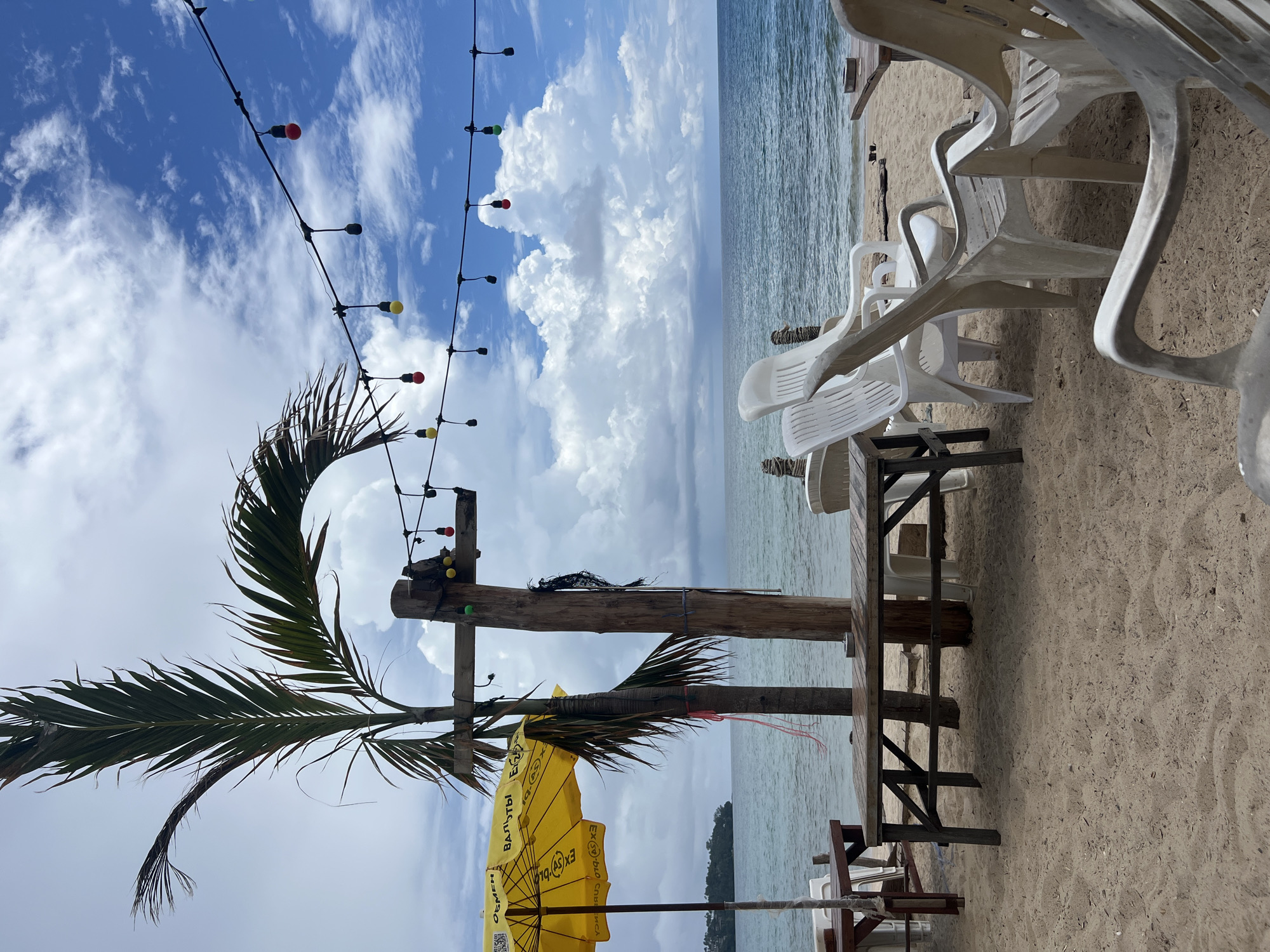 An outdoor beachfront cafe seating area is depicted on a sandy beach in Choeng Thale, Thailand. The scene is set under a partially cloudy sky with a gentle sea breeze evident in the palm frond. In the foreground, several weathered plastic chairs are arranged around wooden tables, some with their surfaces visibly wet. A bright yellow umbrella, displaying Cyrillic text, is partially visible to the left. In the mid-ground, a tall wooden post stands planted in the sand, supporting a string of colorful decorative lights that stretch across the frame. The ocean is calm, with a distant shoreline visible on the left. The overall ambiance suggests a quiet, tropical, and somewhat rustic setting.