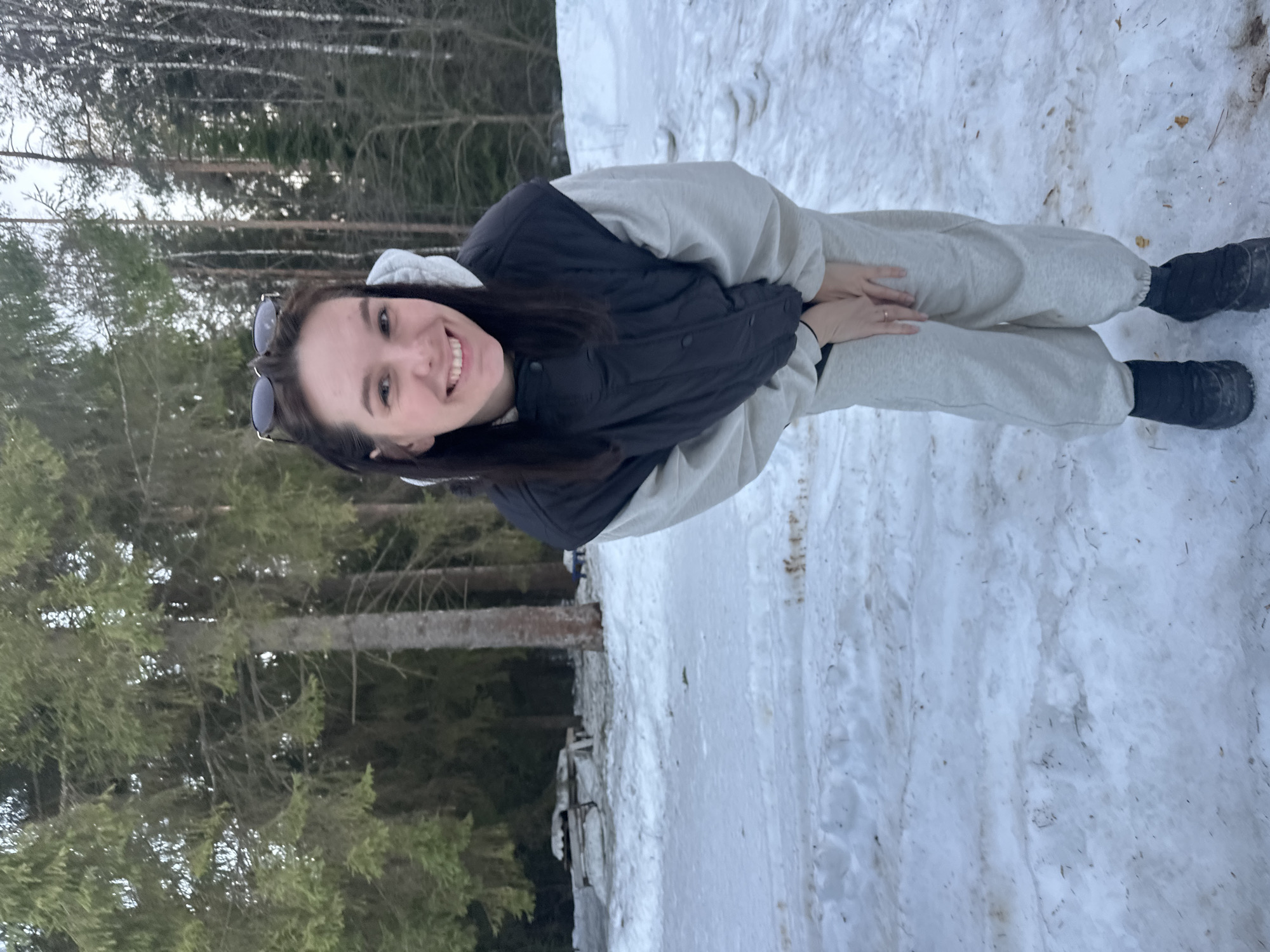 A young female subject is depicted standing outdoors in a snowy environment, identified as Прикамский, Russia. She has dark hair, with dark-lensed sunglasses resting on her head, and is smiling directly at the camera. The subject is wearing a light grey hooded sweatshirt, a dark grey or black puffer vest, light grey sweatpants, and dark-colored winter boots. Her hands are clasped together in front of her waist. The ground is covered in patchy, partially disturbed snow, revealing some underlying dirt and foliage, with visible tracks. The background consists of a dense forest composed of both coniferous and bare deciduous trees. In the left mid-ground, a snow-covered wooden picnic table and bench are visible. A partially obscured blue object is present in the far right background.