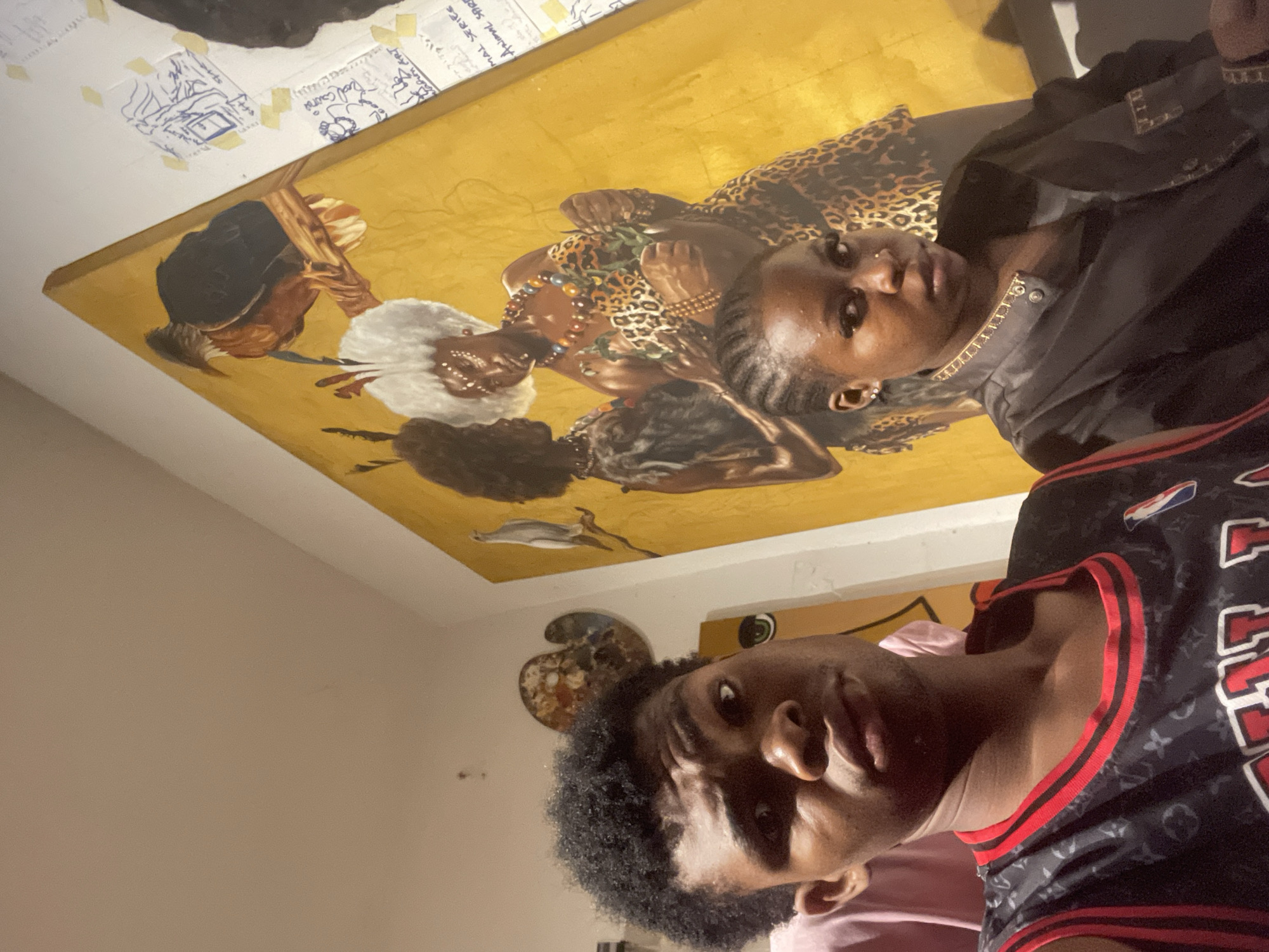 In Ado, Nigeria, a young man and a young woman are photographed in front of a large, vibrant painting. The man, in the foreground and to the left, wears a black basketball jersey with red trim and a subtle Louis Vuitton monogram pattern. He is looking directly at the camera with a neutral expression. To his right and slightly behind him, the young woman is visible, looking towards the camera as well. She has braided hair and is wearing a black jacket with decorative gold detailing.
The painting behind them is a striking piece on a golden background. It depicts two figures, appearing to be women, adorned with traditional jewelry and patterned clothing. One figure has striking white, voluminous hair and is wearing a leopard print outfit and a beaded necklace. The other figure, to the left, has dark, curly hair and is also wearing decorative adornments. Above the figures, on a branch, is a detailed painting of a bird. To the right of the painting, some papers with drawings and text are taped to the wall. The overall environment appears to be an indoor space, possibly an art gallery or studio, with neutral-colored walls.