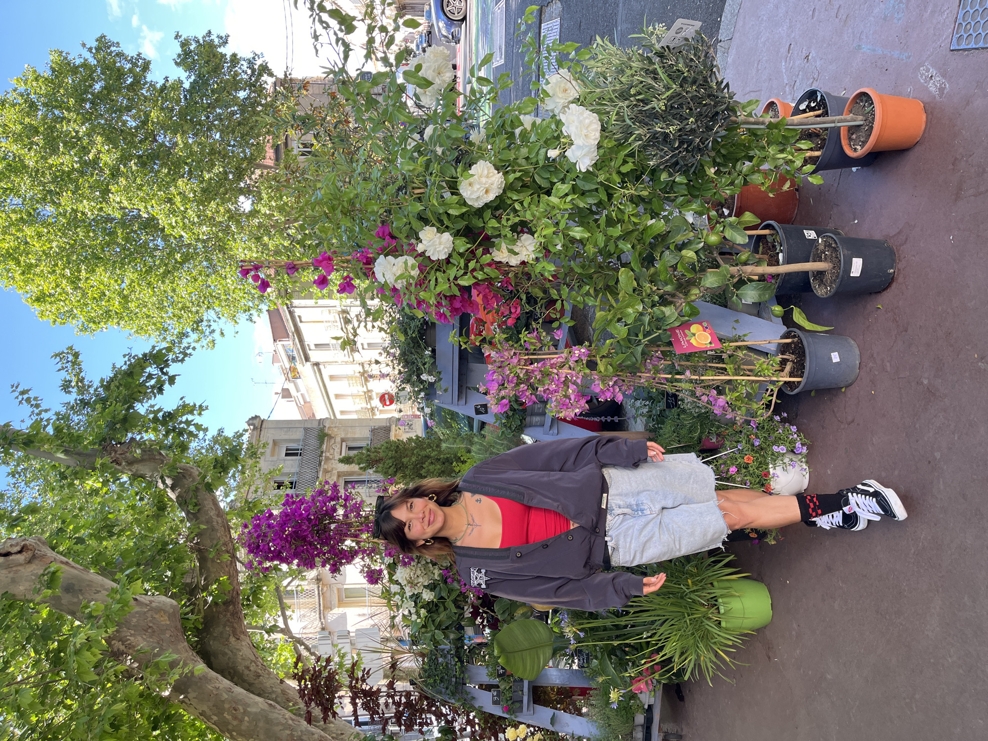 A young woman stands outside a flower shop in Montpellier, France. She is wearing a red tank top, grey jacket, and light blue jean shorts, with black and white sneakers and patterned socks. She is surrounded by plants and flowers, including white roses, bougainvillea, and other potted greenery. The flower shop is located on a street with buildings visible in the background. A large tree with green leaves provides shade on the street.