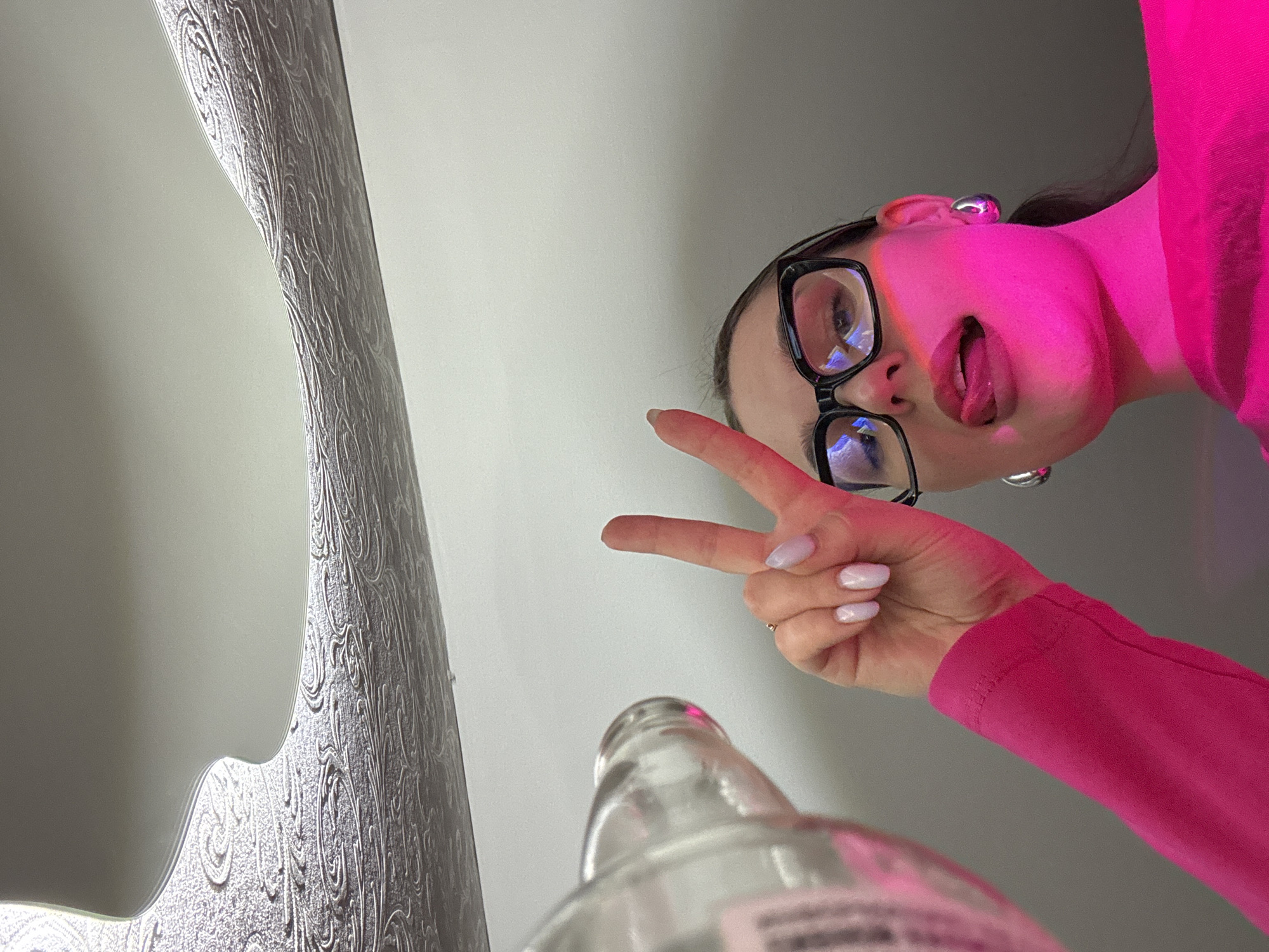 A low-angle, upward-facing shot captures a young person, likely female, peering down into the camera lens. She is wearing large black-framed glasses and prominent silver hoop earrings. Her face and upper body are bathed in a vibrant pink artificial light. She has her mouth slightly open with her tongue sticking out in a playful expression. Her right hand, adorned with light-colored manicured nails, forms a peace sign. She is dressed in a bright pink long-sleeved top. In the immediate foreground, a clear glass bottle, possibly for an alcoholic beverage, is visible but blurred, with an unreadable label on its side. To the left, a textured, light-grey surface with an ornate, flowing pattern is visible, possibly decorative wallpaper or a mirror frame. The background consists of a plain, light-colored wall or ceiling. The image conveys a casual and playful mood, possibly taken during a social event indoors, in Chaikovsky, Russia. The artificial pink lighting suggests it might be evening or night.