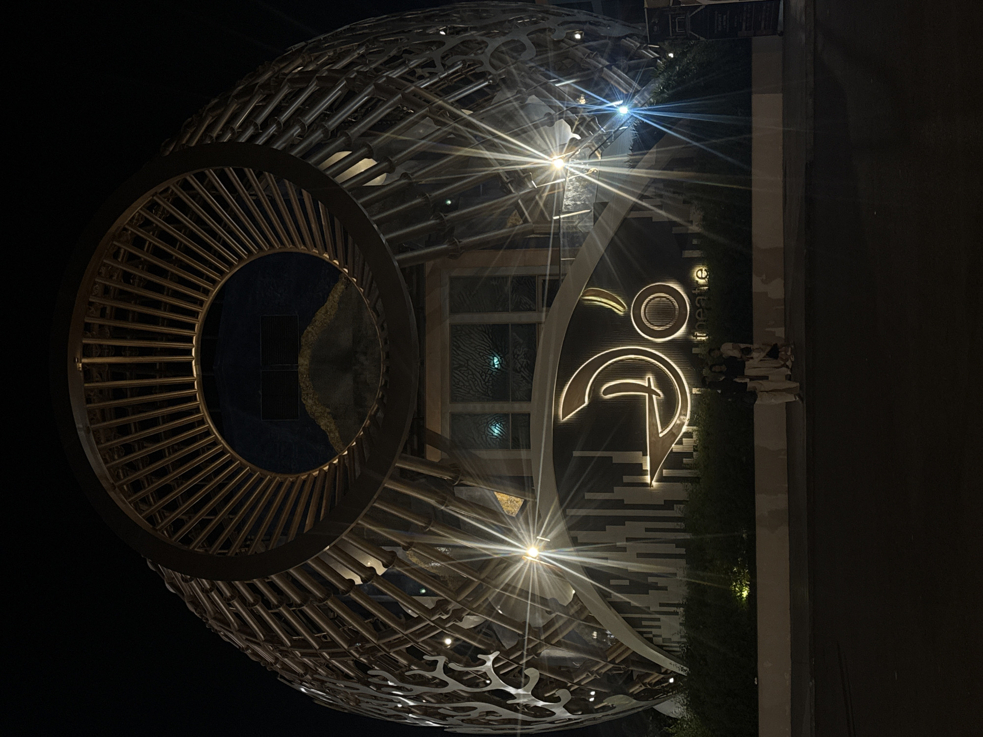 The image depicts the exterior of a large, spherical building at night, illuminated by spotlights that create starburst effects. The building's facade is a complex structure of interwoven metal beams and decorative panels. A prominent glowing logo is visible on the front, with the word "THEATRE" below it in smaller lettering. The location is identified as Nha Trang Ward, Bac Ninh City, Vietnam. In front of the building, a low-lying hedge runs along a white-washed wall. Three people are standing on the sidewalk in front of the hedge, facing the building. The ground in front of them is asphalt. The sky is dark and black, indicating nighttime.