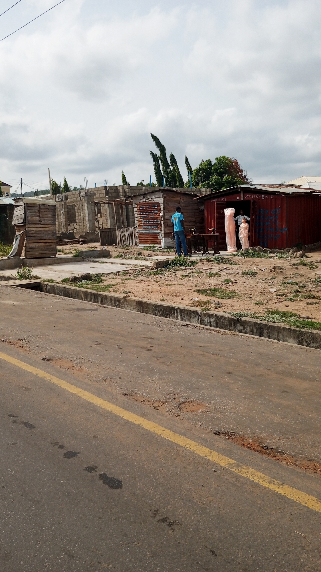 The image shows a street scene in Zango-ndaji, Nigeria. The road, marked with a yellow line, is cracked and shows signs of wear. A concrete curb separates the road from a dirt area containing sparse vegetation. Several informal structures are visible: a half-constructed building of gray bricks, two wooden shacks, and a red shipping container. A man in a blue shirt stands near a sewing machine outside the shacks. A dressmaker's dummy wrapped in plastic stands in the doorway of the red container. Trees and foliage can be seen in the background. The sky is overcast.