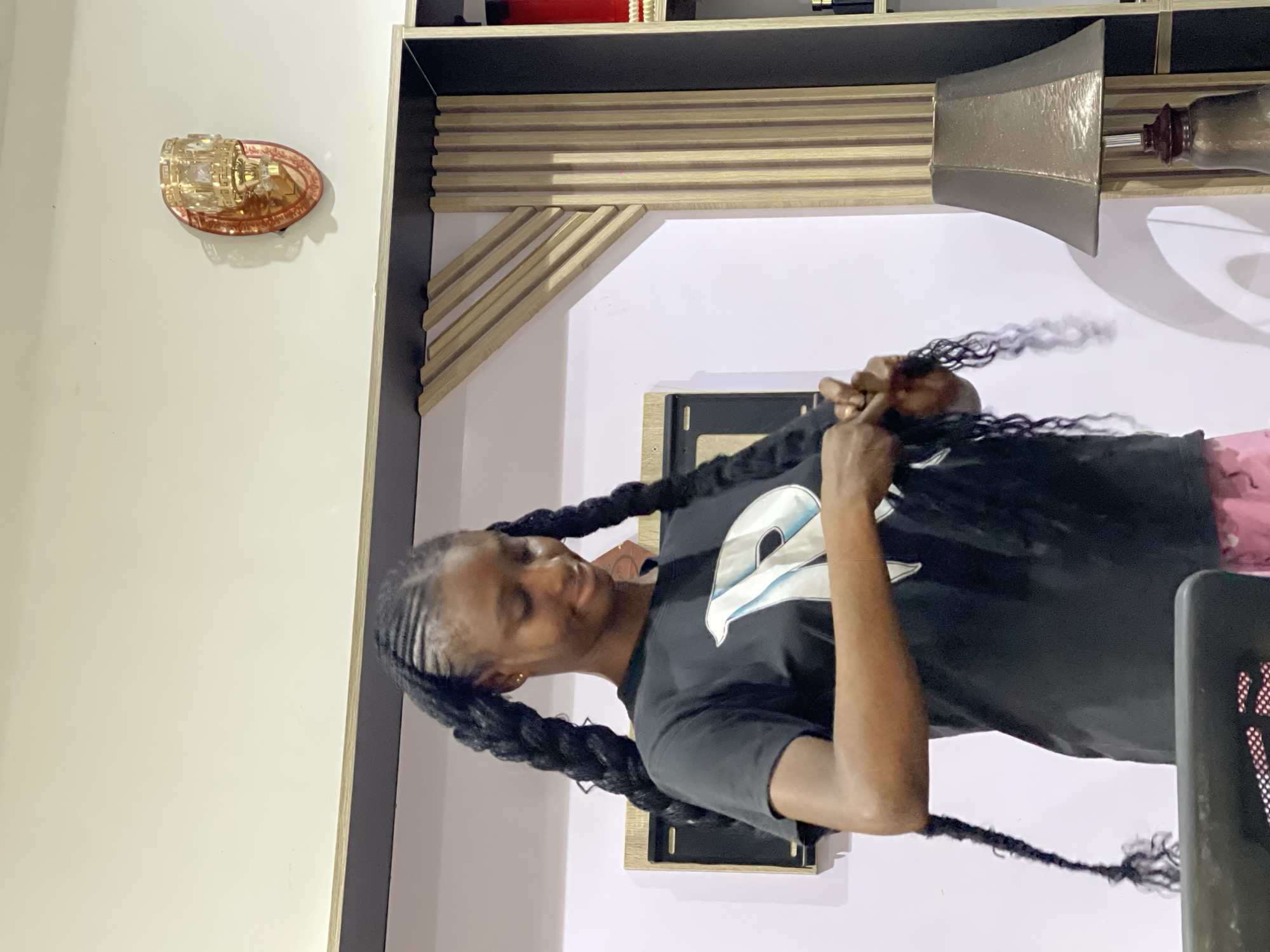 The image captures a young Black woman with dark skin and braided hair in two large pigtails, standing indoors and appearing to be doing her hair. She is wearing a black t-shirt with a white logo on the front and pink shorts. She is looking down and her hands are engaged with her hair, suggesting she is in the process of styling it.
The background suggests an indoor setting, possibly a home or salon. The walls are white, and there's a decorative light fixture on the wall to the right, which appears to be made of gold-toned metal and glass. A wooden shelf unit with vertical slatted panels is visible behind the woman, and a lamp with a textured, dark lampshade sits on a surface to the right. There's also a framed picture or decorative item on the wall behind the woman.
Given the prompt specifies the location of Abuja, Nigeria, this scene is situated within that city. The overall environment appears to be a well-lit domestic or commercial space.