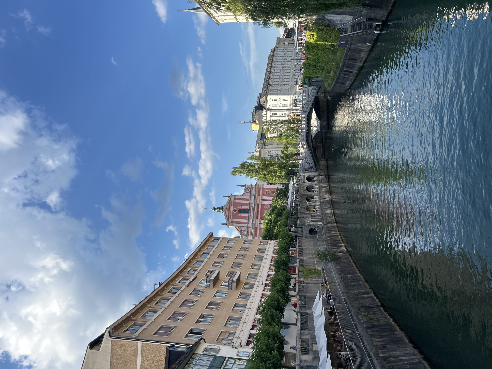 This is a vertical, eye-level, wide-angle photograph of a canal or river in a European city. The water is a deep blue-green and reflects the sky and surrounding buildings. Along the bank, there is a stone walkway lined with trees and buildings. On the left side of the canal, a multi-story building with many windows is visible. Further back, a pink church with a green dome stands out against the sky. On the right side of the canal, more buildings with white facades and ornate architectural details are visible, as well as a bridge spanning the water. The sky is a clear blue with scattered white clouds. People can be seen walking along the canal bank and sitting at outdoor cafes. The sun is shining, casting reflections on the water, suggesting it is daytime. The scene is bustling and vibrant, indicating a pleasant day for outdoor activities in a city known for its historic architecture and waterways. The overall mood is one of peaceful activity and urban charm.