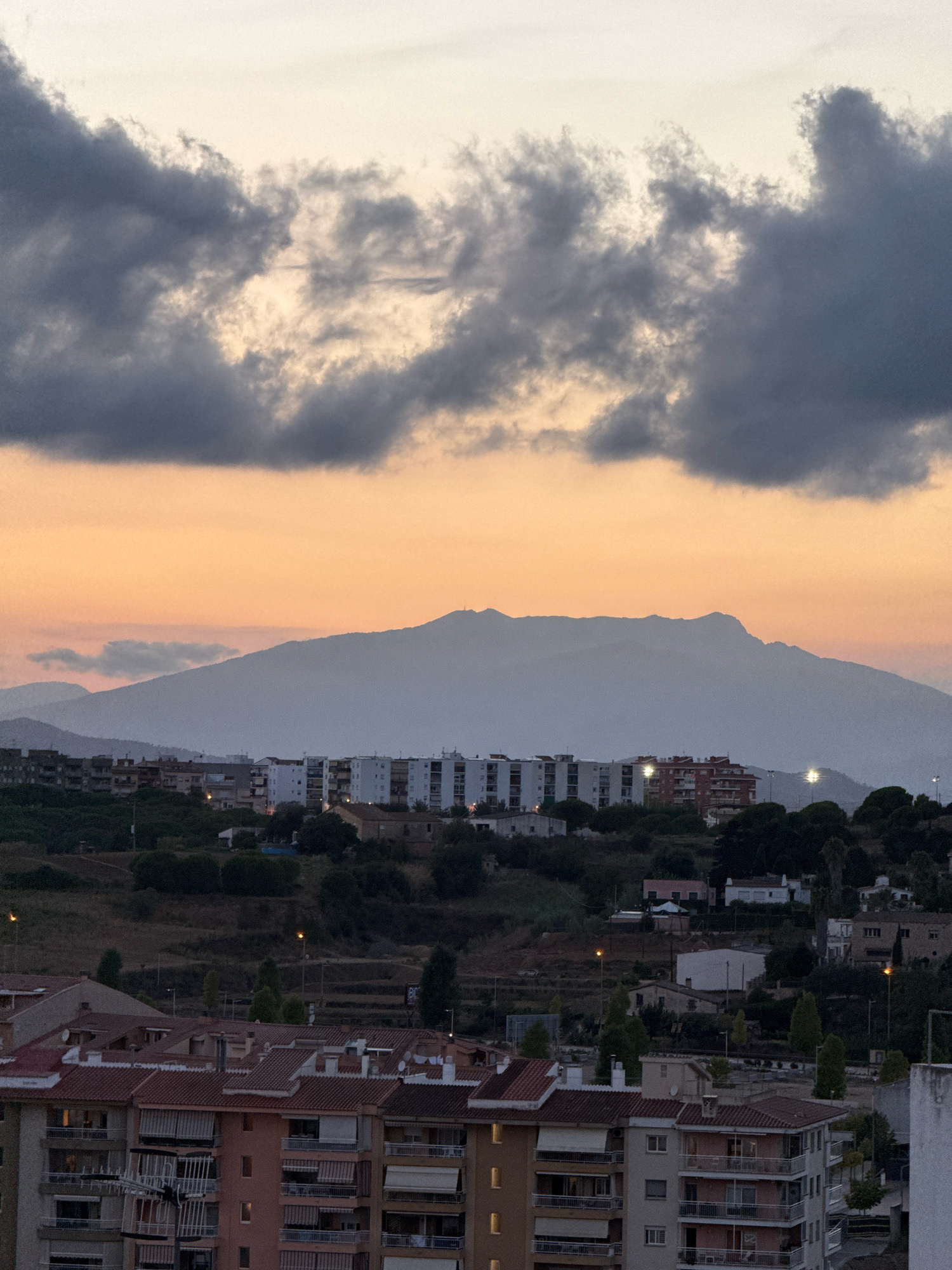 The image depicts a twilight scene over the city of Blanes, Spain. A dramatic sky with dark, heavy clouds contrasts with a soft, orange glow near the horizon, hinting at the setting sun. In the mid-ground, a range of hazy, blue mountains forms a silhouette against the sky. Below the mountains, a low-rise urban landscape unfolds. Buildings, predominantly residential blocks with white and light-colored facades, are visible. Some buildings have balconies and awnings. Trees and vegetation are interspersed throughout the cityscape and on the rolling hills behind it. The overall atmosphere is serene and peaceful, characteristic of dusk in a coastal town.