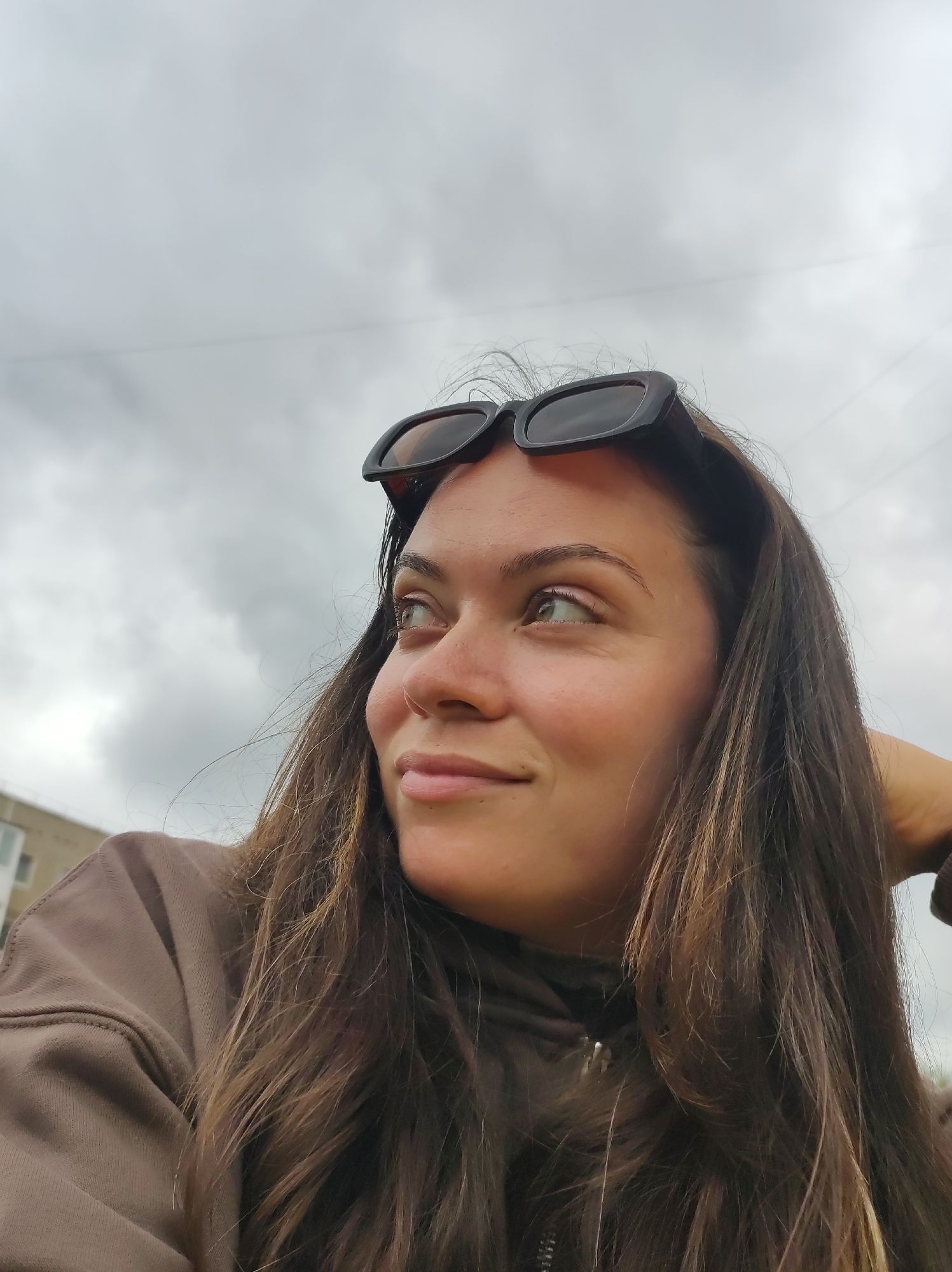 A close-up, waist-up, outdoor portrait shows a young woman with long, dark brown hair wearing sunglasses on top of her head and a brown hooded sweatshirt. She is looking to her left with a slight smile. Her right hand is visible behind her head. In the background, a building and a cloudy sky are visible. The photo was taken in the city of Chaikovsky, Russia.