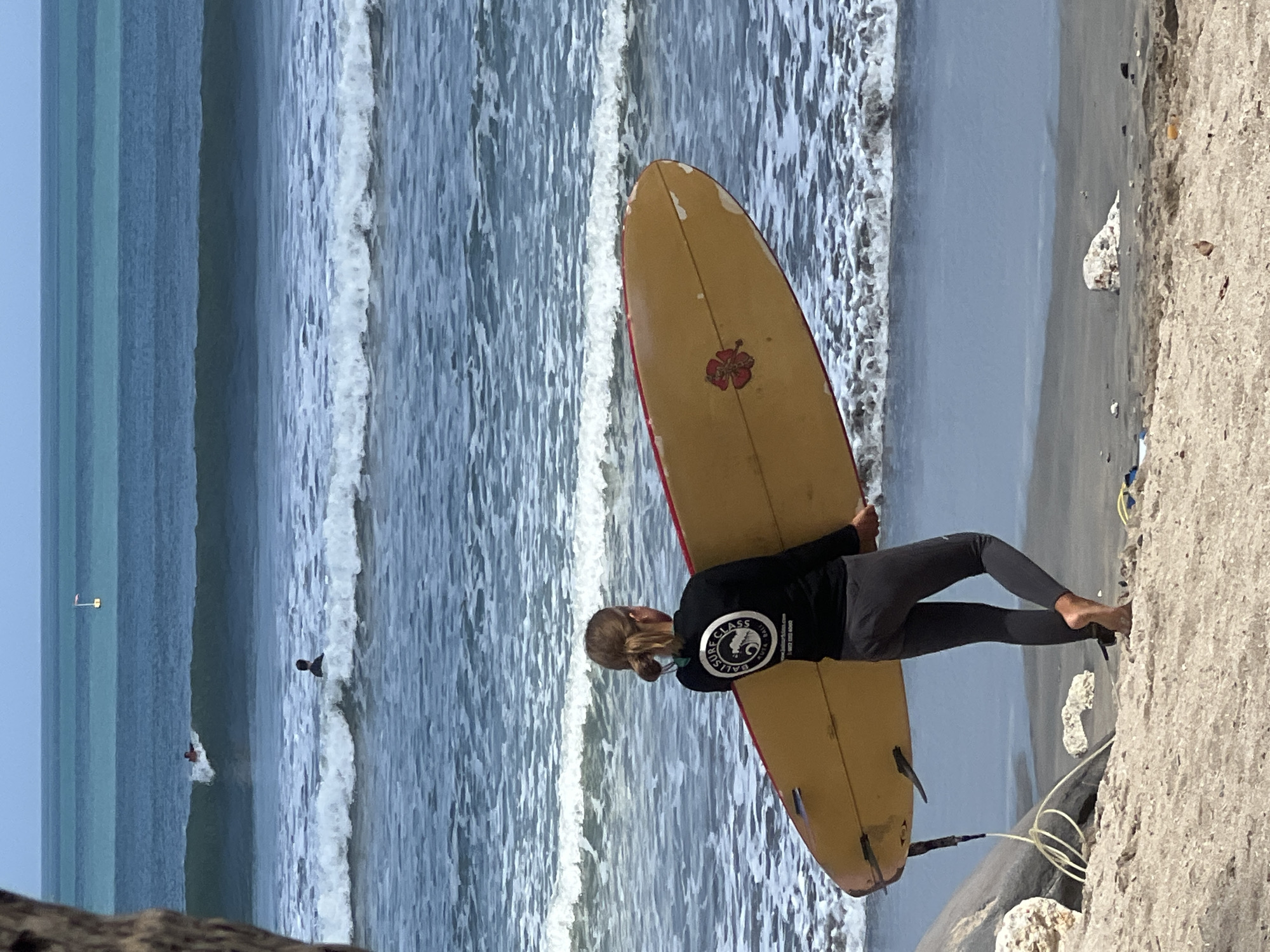 An eye-level, medium shot captures a young woman on a beach in Kuta, Indonesia, holding a yellow surfboard. She is positioned in the foreground, facing away from the camera and towards the ocean. Her dark long-sleeved shirt features a circular logo with text and an image of a hibiscus flower. She wears dark leggings that extend down to her ankles, revealing her bare feet in the sand. The surfboard, with a red trim and a red hibiscus design on its surface, rests under her left arm and is angled towards the water.
The beach is composed of light-colored sand, with scattered rocks and debris. To the left of the woman, a large, rough-textured rock is partially visible. The ocean stretches out behind her, with distinct lines of waves. In the middle ground, two surfers are visible in the water. One is actively riding a wave, while the other is paddling. In the distance, the ocean appears calmer with multiple horizontal bands of varying blue and turquoise hues, suggesting different depths or water conditions. A small white flag on a pole is visible on the horizon in the far distance. The sky is a pale blue.
