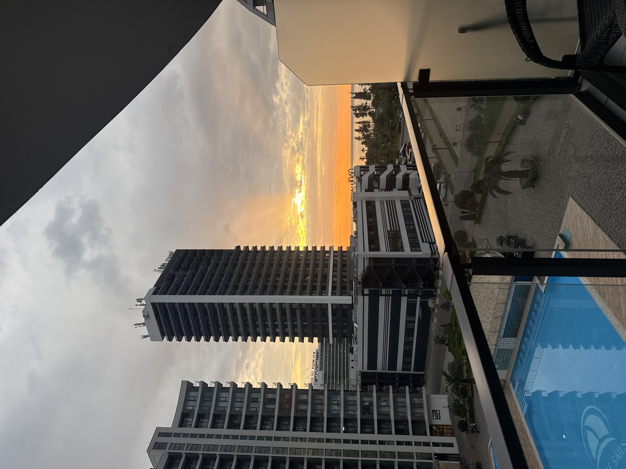 The image shows a cityscape at dusk, likely in Batumi, Georgia, due to the modern architecture and the proximity to the coast. From the vantage point of a high balcony, a few buildings are centered in the frame.
The main building is a tall, modern tower with horizontal bands of windows running its length. To the side, there is another building with similar architectural elements, though shorter.
Between the buildings, the setting sun is visible, casting an orange glow over the landscape and reflecting on the overcast sky. The Black Sea is visible in the distance.
On the right, the balcony of the building where the photo was taken has a glass railing. Below, there is a swimming pool and paved area with greenery. There are some plants and trees, along with signs of infrastructure like roads and paths.
The environment suggests an urban location with modern amenities and coastal proximity.