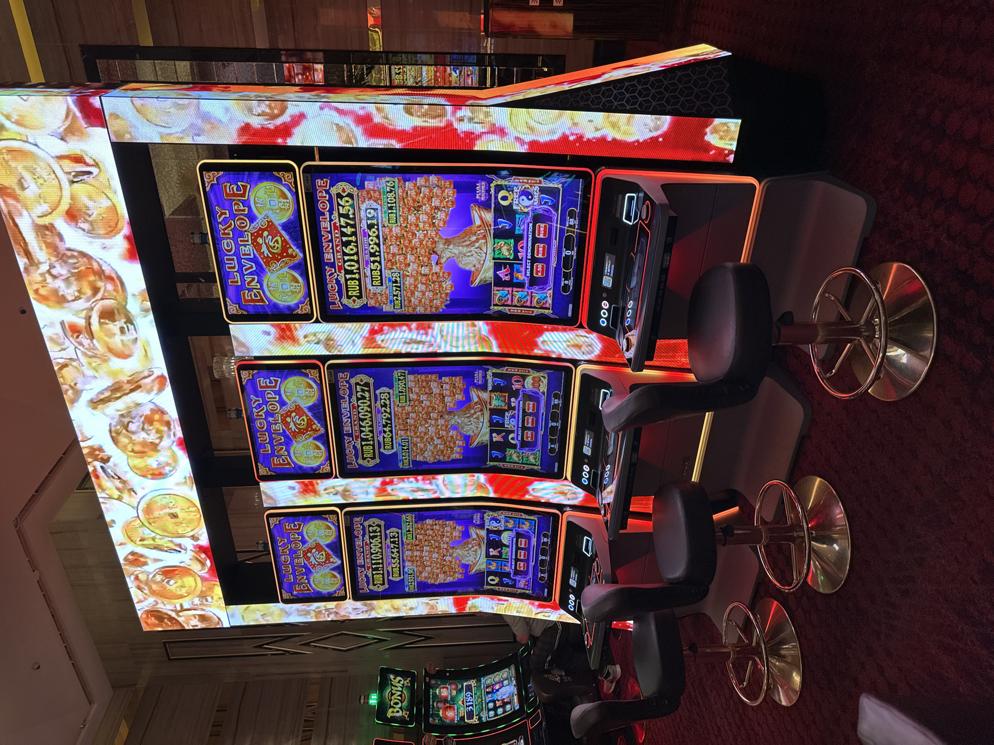 The image shows a row of three modern slot machines with large, brightly lit LED screens displaying graphics and game information, specifically highlighting the game "Lucky Envelope" with large jackpot amounts. Two slot machines on the right are identical, featuring a tree with red envelopes and large jackpot figures in Russian Rubles. The slot machine on the left is slightly different, showing "Bonus" and a different game interface with a number "3189". In front of the slot machines are three black bar stools with gold bases, arranged for players. The floor is covered in a textured red carpet. The background shows the interior of what appears to be a casino, with subtle geometric patterns on a wall to the right and the edge of another slot machine visible to the far left. The lighting is dramatic, with the slot machines and their displays casting a red and orange glow. The scene suggests a gambling establishment in Estosadok, Russia.