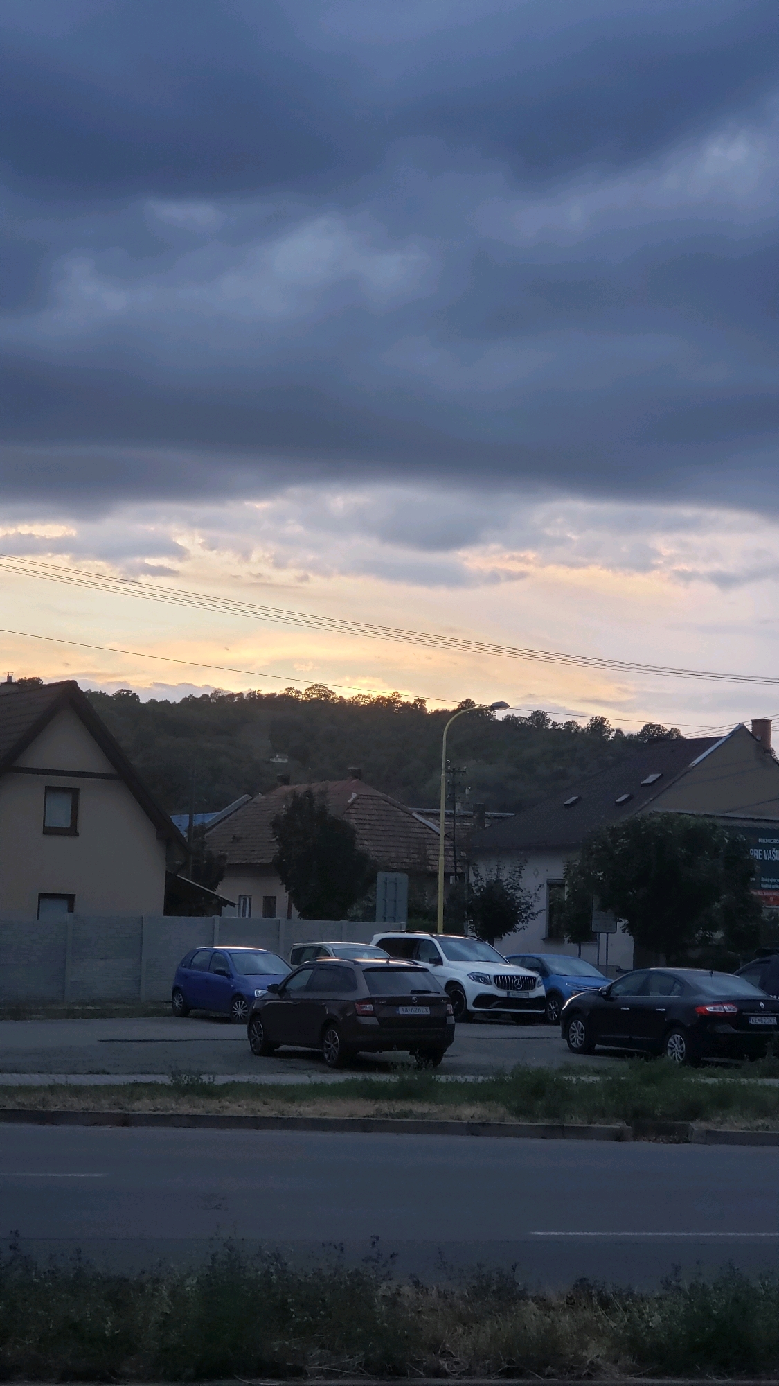 A twilight scene unfolds in the city of Prešov, Slovakia, with a brooding, dark cloud cover dominating the upper portion of the image. A sliver of pale yellow light breaks through the clouds near the horizon, illuminating the silhouettes of trees and the crest of a densely wooded hill. Below the sky, a residential area is visible, characterized by houses with tiled roofs and neutral-colored facades. Power lines stretch horizontally across the frame, bisecting the sky and the landscape. In the foreground, a row of parked cars is visible on asphalt. Among them are a dark station wagon, a blue hatchback, a white SUV, and a dark sedan. The street is paved, with a strip of dry grass and weeds lining the edge. The overall atmosphere is subdued, marked by the fading light and the impending weather.