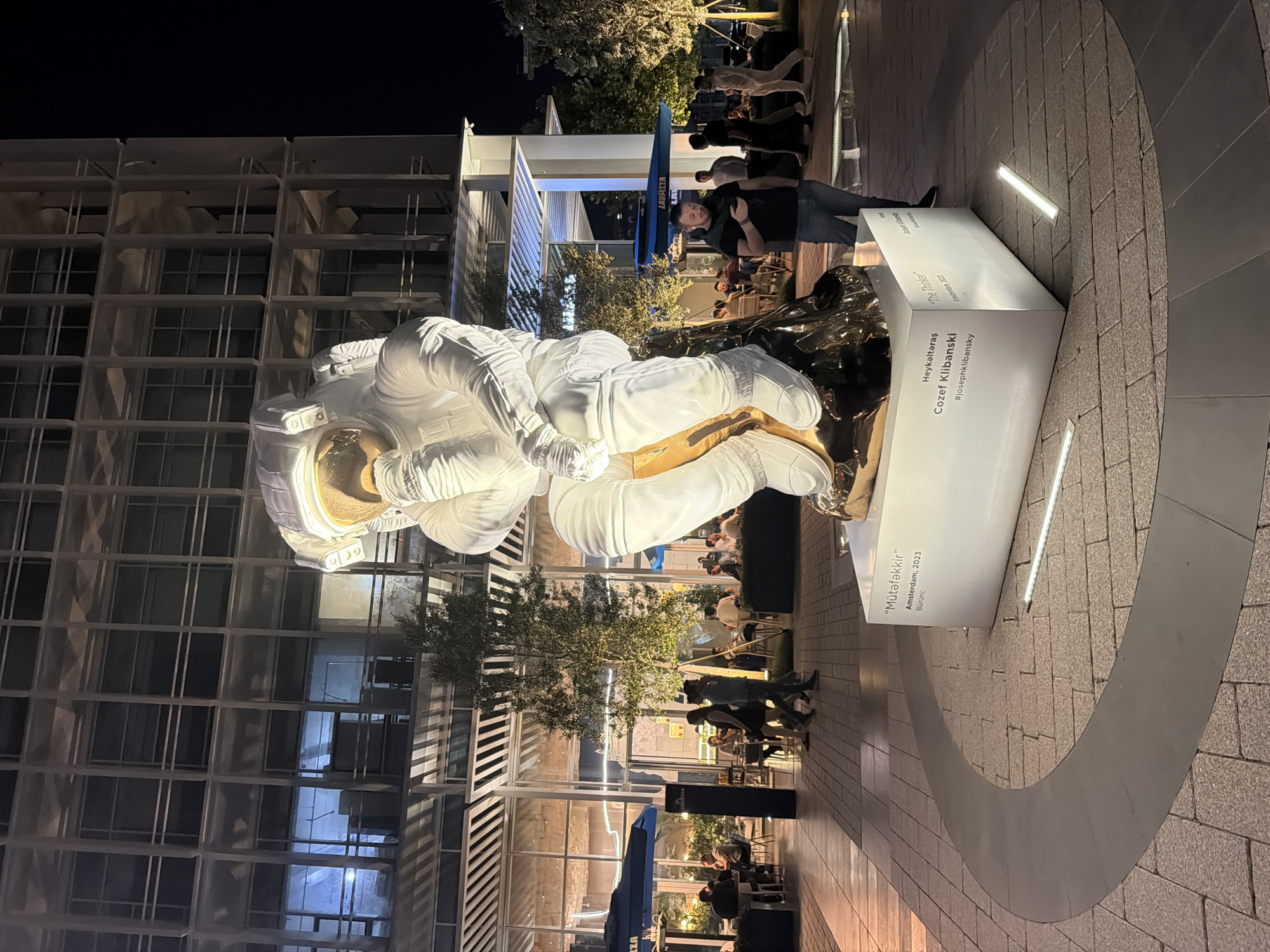 The image shows a large sculpture of an astronaut in a contemplative pose, located in an outdoor plaza at night. The sculpture is predominantly white with gold accents on the helmet visor and parts of the body. It is mounted on a white pedestal that displays text, including the title "Mütafakkir" and "The Thinker," along with the artist's name, Cozef Klibanski, and the location "Amsterdam, 2023". The background features a modern building with a grid-like facade and large windows, some of which are illuminated from within. Several people are visible in the plaza, some seated at outdoor tables in a restaurant area under umbrellas, while others are walking or standing. The ground is paved with stone tiles arranged in a circular pattern around the sculpture. The overall lighting suggests an urban environment at night. The location is specified as Khatay Raion, Azerbaijan, though the text on the pedestal indicates Amsterdam, 2023.