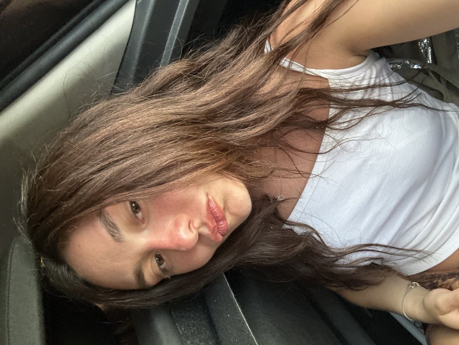 An indoor, close-up, front-facing image features a single individual, a female, seated within the interior of a vehicle. Her long, dark brown hair cascades over her shoulders and chest. Her face exhibits a reddish hue on the nose and cheeks, consistent with a sunburn. Her lips are pursed in a pout, and her gaze is directed towards the camera. She wears a white, thin-strapped tank top, a delicate chain necklace with a small circular pendant, and a light-colored bracelet on her left wrist. A small portion of an animal print garment is visible beneath her white top. The background consists of dark, indistinct elements of the vehicle's interior, including a dark seat or headrest to her left and a car door frame to her right. The specific location within the city of Deià, Spain, is not discernible from the provided visual information.