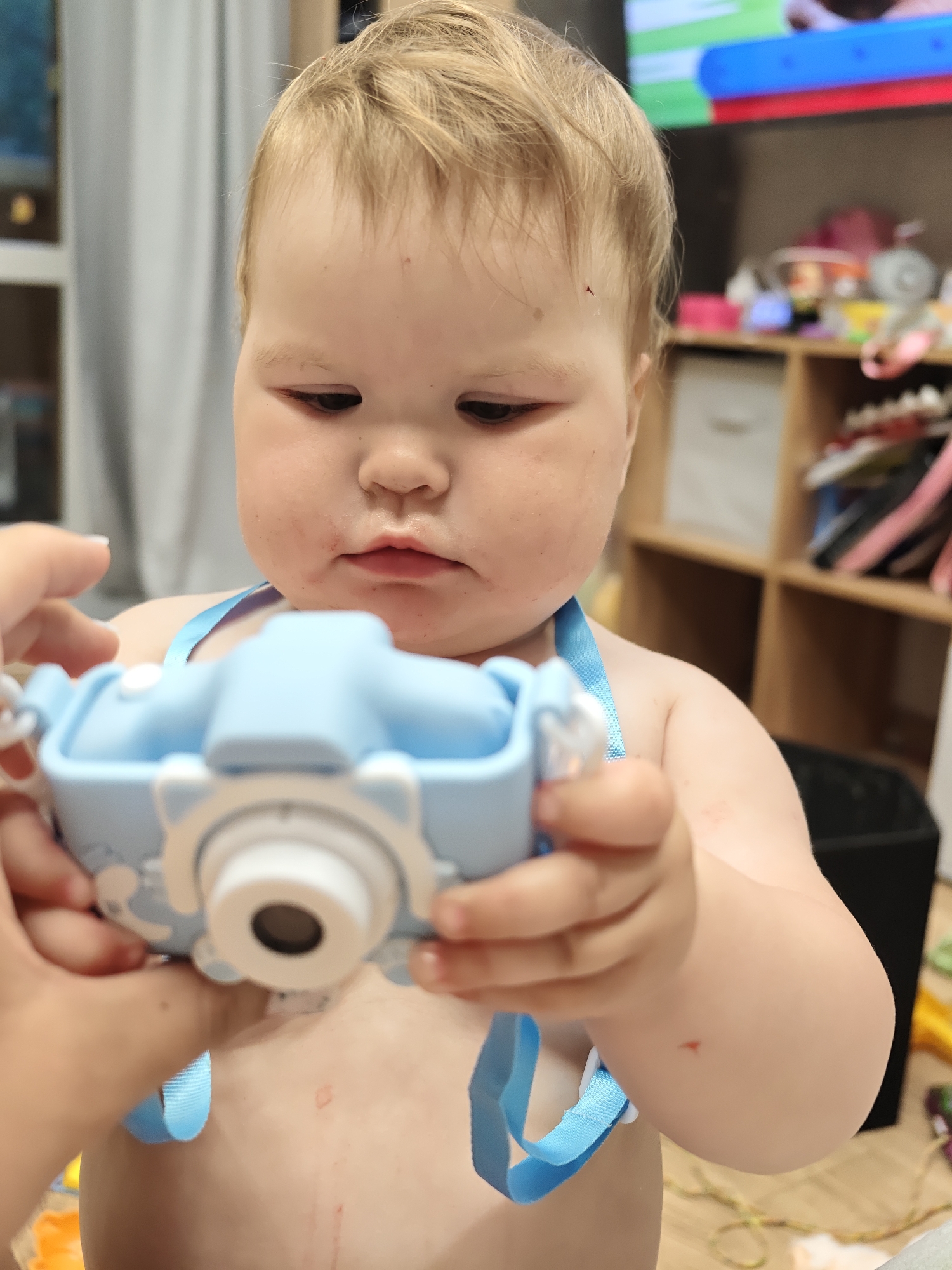 A toddler with blonde hair and fair skin holds a light blue toy camera with a white lens in both hands. The camera has a blue strap around the toddler's neck. The toddler's face and chest have some red marks. A person's hands are visible on the left, assisting with the camera. The background is a cluttered room with a television, a shelf filled with toys and miscellaneous items, and a dark-colored wastebasket. The location is in the city of Chaikovsky, Russia.