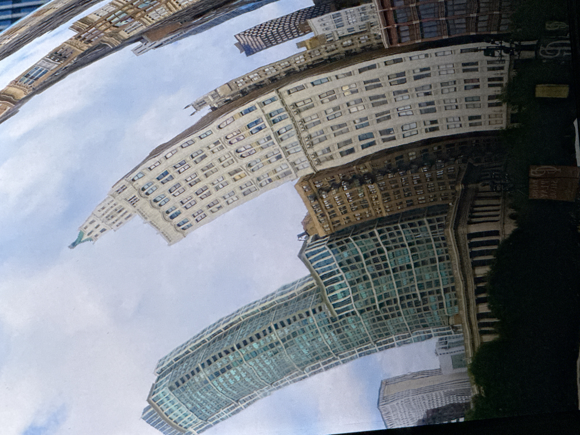 The image depicts a distorted reflection of a city skyline, with multiple skyscrapers of varying architectural styles. The buildings appear warped and stretched, suggesting the reflection is from a curved or uneven surface. The sky is visible with scattered clouds. The scene is likely a photograph of a reflective sculpture or a building with a highly polished, convex facade, creating a fisheye lens effect on the surrounding urban environment. The reflection captures tall buildings made of glass, brick, and stone, with numerous windows and distinct rooflines. The angle of the shot is from a low perspective, looking upwards. The provided context of Saratov, Russia, suggests that this reflective structure is situated in that city, presenting a unique perspective of its urban landscape. No people or specific actions are discernible.