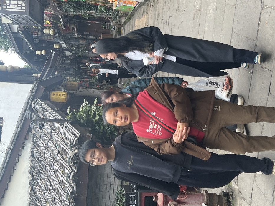 The scene depicts a pedestrian street within Ciqikou Ancient Town, Shapingba District, Chongqing, China. The environment features traditional Chinese architecture with tiled roofs and wooden structures, numerous yellow and red lanterns, and dense foliage, indicating an old town or historical commercial area.
In the foreground, three individuals of East Asian descent are visible. On the left, a young male with short dark hair and glasses wears a dark blue long-sleeved shirt with "TREND RIDER" text, an open dark hoodie, and dark pants. He is looking towards the right. In the center, a middle-aged female with dark hair pulled back wears a red long-sleeved shirt with white text, a brown open jacket, brown pants, and a black shoulder bag with a red and green strap. Her hands are clasped in front of her, and she is looking directly forward. On the right, a young female with long dark hair and glasses wears a white long-sleeved shirt, a long dark grey coat, dark pants, and light-colored sneakers with blue accents. She holds a white-patterned disposable cup in her right hand and a clear plastic bag containing unspecified items in her left, looking towards the left.
Behind these individuals, another female in a green jacket is partially visible. Further down the narrow street, several other pedestrians are present, walking in both directions. A red fire hydrant is visible on the far left. Overhead, multiple yellow lanterns are suspended, one clearly displaying Chinese characters "老磁器" (Lao Ci Qi - Old Ciqi). The street is paved with grey stones, and the lighting suggests an overcast sky.