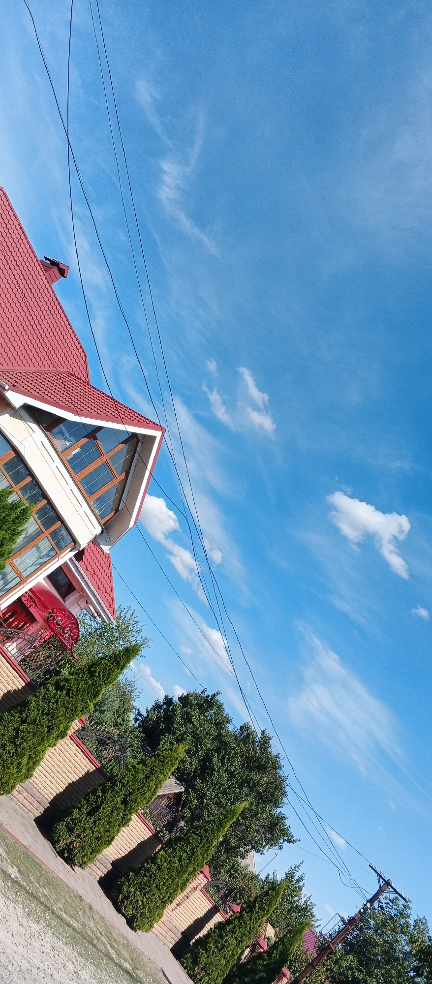 The image depicts a section of a residential property in Smila, Ukraine. The foreground shows a paved walkway and a fence constructed from light brown brick with a red trim. Along the fence, several tall, conical evergreen trees are planted, casting shadows. Behind the fence, dense green trees form a backdrop. A portion of a two-story house is visible on the left side of the frame, featuring a red tiled roof, cream-colored siding, and large windows with wooden frames. A red ornamental awning is above a doorway on the lower level of the house. The sky above is a clear blue, with scattered white wispy clouds and several black utility wires crisscrossing the frame. The image is taken from a low angle, and the scene is tilted.