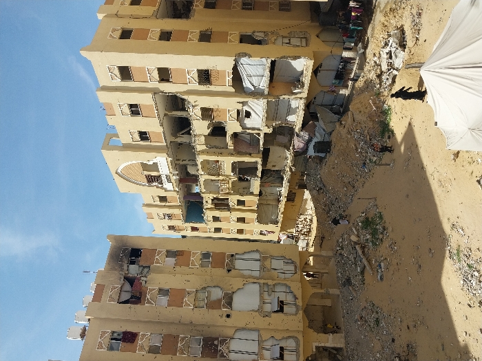 The image captures a scene of severe structural damage in Khan Yunis, Palestinian Territory, under a clear blue sky. Two multi-story, light-colored buildings stand prominently, both exhibiting extensive damage. The building on the left shows numerous impact marks, possibly bullet holes, across its facade, with broken window frames and scorched areas, particularly on the upper floors. White water tanks are visible on its roof. The building on the right has significant sections of its central facade and internal floors collapsed, exposing rebar and debris, while also displaying similar impact marks and broken windows. Some openings in this building are covered with white tarps or makeshift materials, and clothing hangs on lines near its lower right section.
The ground between and in front of the buildings is largely sandy, interspersed with numerous piles of rubble, broken concrete, and shattered building materials. Several dirt paths traverse this debris-strewn ground. Multiple individuals are present: one person in dark clothing walks across the sandy foreground from left to right; two individuals, one in white and one in dark attire, walk on a path further back near the center; and additional figures are visible near the bases of both damaged buildings. A large white tent or tarp structure is partially visible in the bottom right foreground. The overall environment conveys a scene of significant destruction, yet with evidence of human activity.
