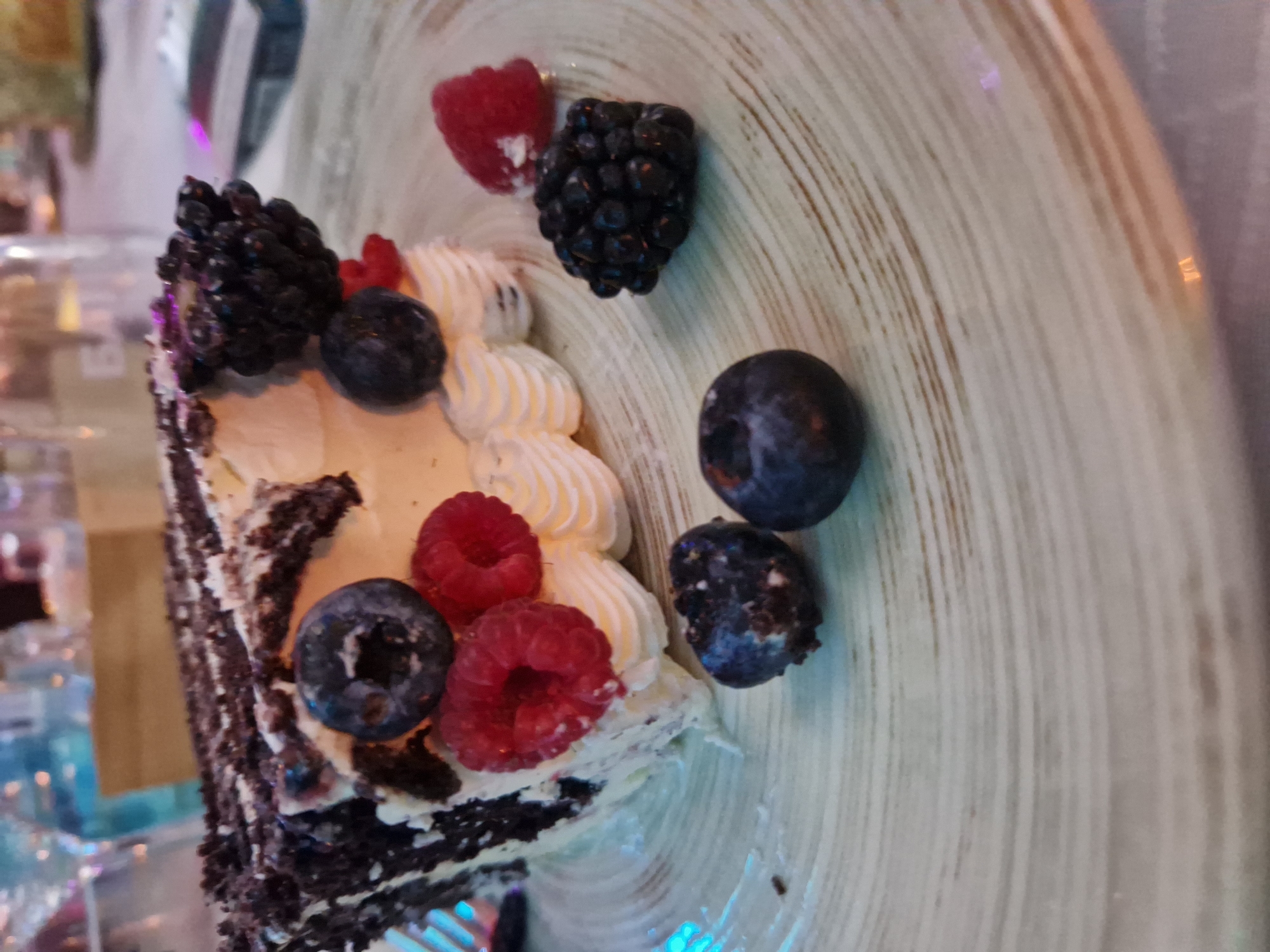 A slice of chocolate cake is on a plate. The cake has layers of dark chocolate cake and white cream. It is topped with dark chocolate shavings, raspberries, blueberries, and blackberries. The plate is light-colored with concentric rings. In the background, out of focus, are bottles and other tableware. This image was taken in Tankovo, Bulgaria.