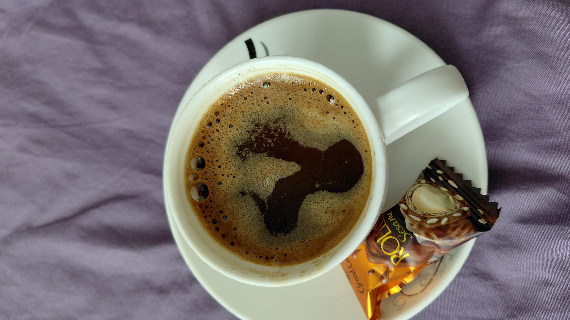 A close-up, overhead shot shows a white ceramic cup of coffee sitting on a white saucer, which is placed on a wrinkled lavender fabric. The coffee is dark brown and topped with a layer of foam, with several bubbles visible. Within the foam, a dark silhouette appears, resembling a rabbit or bunny shape. To the left of the coffee cup, on the saucer, is a wrapped chocolate candy. The candy wrapper is gold and orange with brown accents, and the text "ROLL" is prominently visible, along with the word "Sesame". The fabric beneath the saucer has visible folds and creases, suggesting it might be a tablecloth or bedding. The overall scene is one of a beverage and a sweet treat, presented in a simple, domestic setting. There is no indication of the location in Abovyan, Armenia.