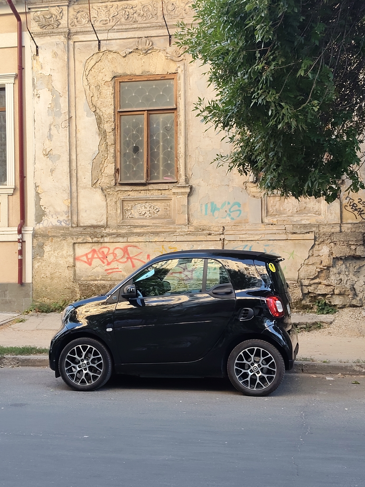 A black Smart Fortwo car is parked on the side of a street in front of an old, dilapidated building. The building's facade is weathered and peeling, with visible signs of decay and graffiti. A tree with dense green foliage hangs over the right side of the scene. The street is paved, and there is a sidewalk in front of the building. The car is positioned parallel to the curb, and its doors are closed. The background shows architectural details of the building, including a window with intricate framing and a decorative stone relief. The overall impression is one of urban decay juxtaposed with the modern car. The car has a yellow sticker with a black exclamation mark on its rear window.