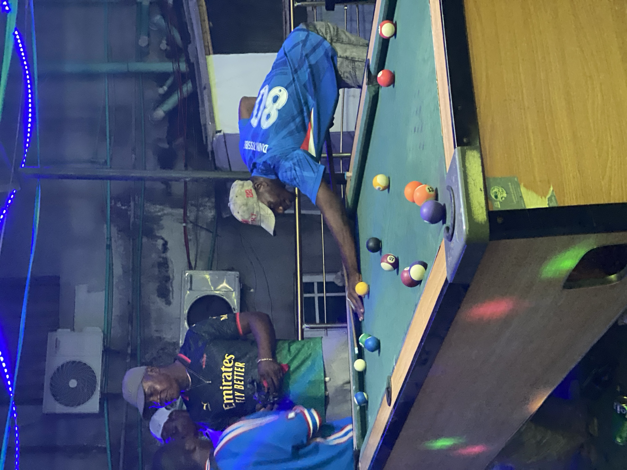 The scene is a nighttime shot of a group of men playing pool indoors, possibly in a bar or recreational space within the city of Ado, Nigeria. One man, wearing a blue jersey with the number 80, leans over a pool table, presumably lining up a shot. Another man, wearing a black "Emirates FLY BETTER" shirt, stands near the table. A third man can be partially seen. The pool table is set with a variety of balls. In the background, there is an air conditioning unit mounted on the wall and blue decorative lights. Metal pipes and beams are visible near the ceiling, suggesting an industrial or unfinished aesthetic.