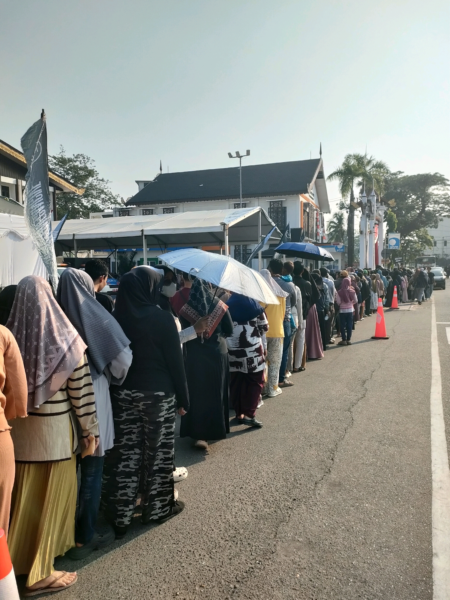 A long line of people, predominantly women wearing hijabs, are standing on an asphalt road in Banjarmasin, Indonesia. The people are facing towards the right side of the frame, suggesting they are queuing for something. Some individuals are holding umbrellas for shade from the sun. In the background, there are buildings with dark roofs, palm trees, and streetlights. The sky is clear and bright. The scene appears to be outdoors during daylight hours.