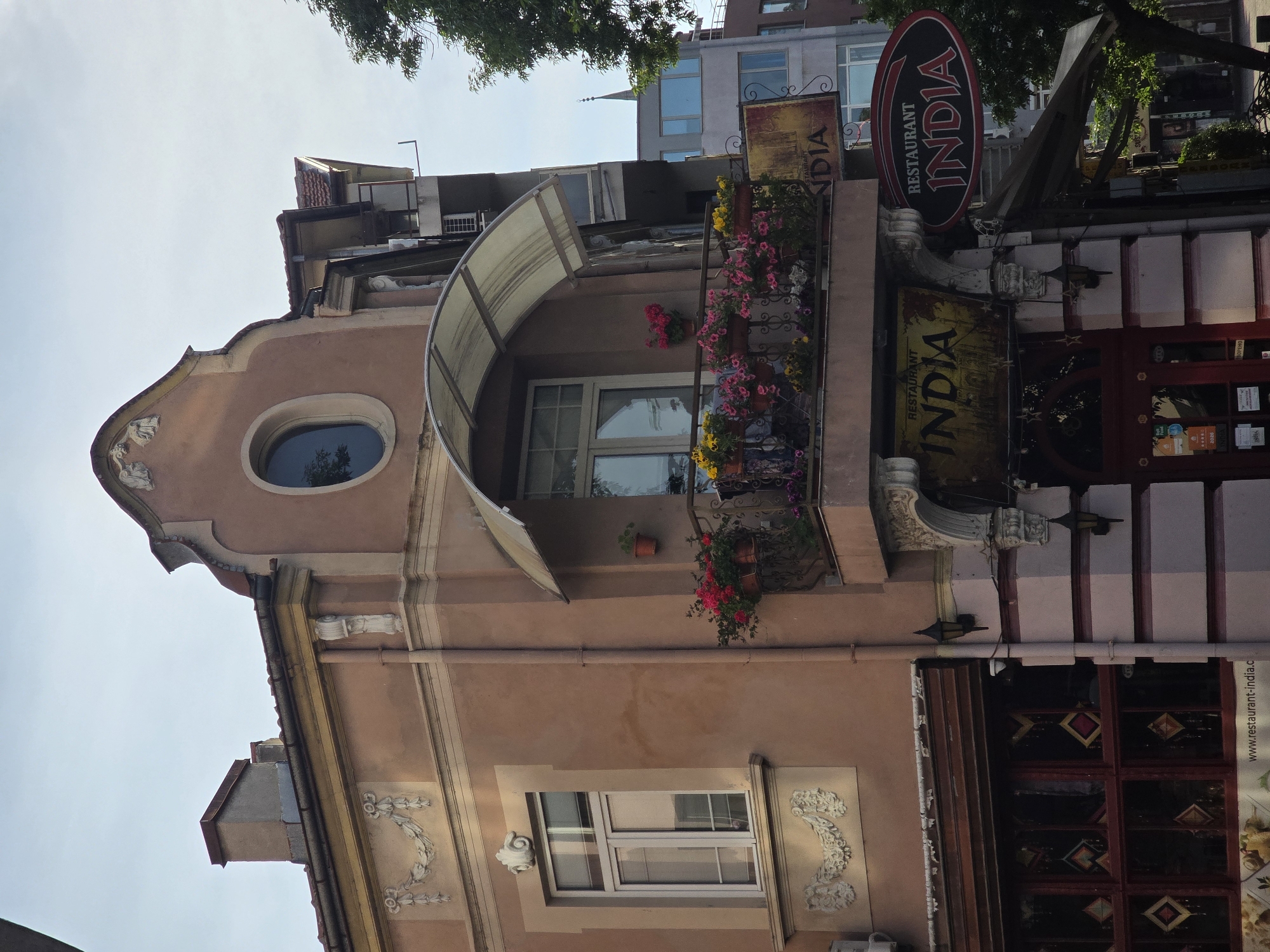 Here is a description of the image:
The image shows the exterior of a building, likely a restaurant, in an unknown city. The building has a peach-colored facade with ornate white trim and a window with a rounded top on the upper level. There is a balcony with potted plants on the second floor and a retractable awning above it. To the right of the building is the entrance to a restaurant named "India," with signage and decorative elements. The image is taken at an upward angle, showing the building against a blue sky and the tops of trees.