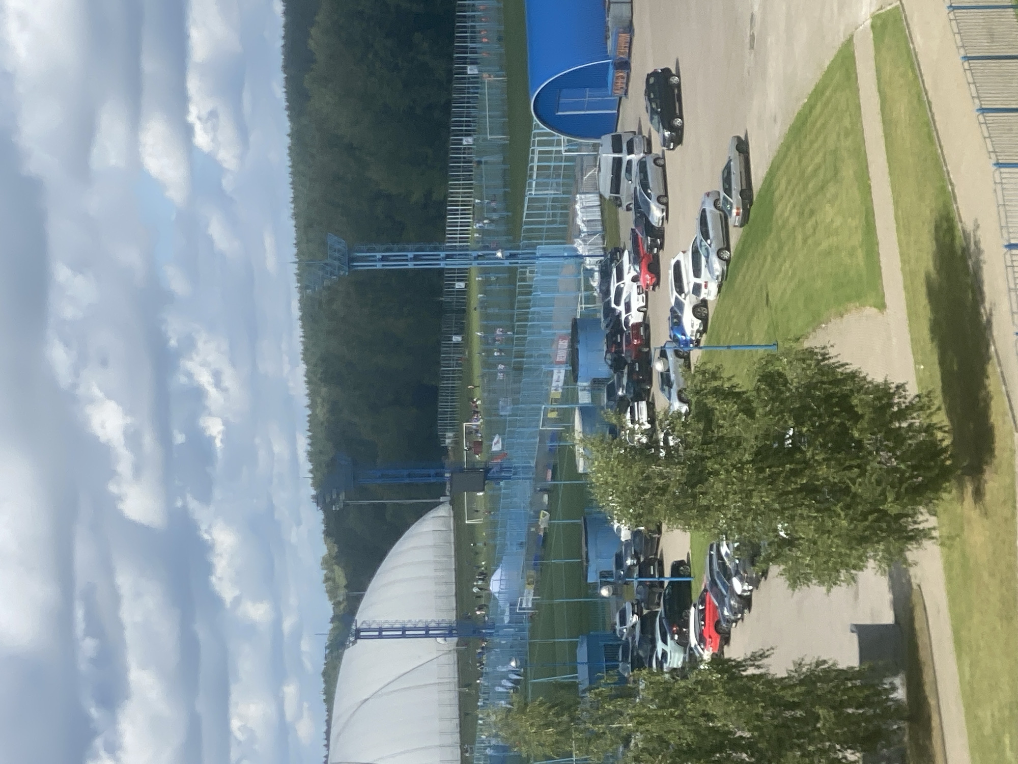 The image captures an outdoor scene at a sports complex on a partly cloudy day. In the foreground, a parking lot is filled with numerous cars of various makes and models, mostly sedans and SUVs. Two large trees with lush green foliage are prominently featured on the left and right sides of the image, casting shadows on the grass and pavement.
Behind the parking lot, a blue fence encloses a green grassy area that appears to be part of a sports field. Beyond the fence, a large white dome-shaped structure, likely an indoor sports facility, is visible. Several tall blue light towers are positioned around the complex, illuminating the fields. Further in the background, a dense forest forms a natural backdrop to the scene. There are small groups of people scattered across the sports fields, suggesting some activity is taking place. A blue arched building is situated to the right of the parking lot. The overall impression is of a functional sports venue with active parking and fields. The location is confirmed to be Minsk, Belarus, specifically at the Minsk Arena ice hockey stadium.
