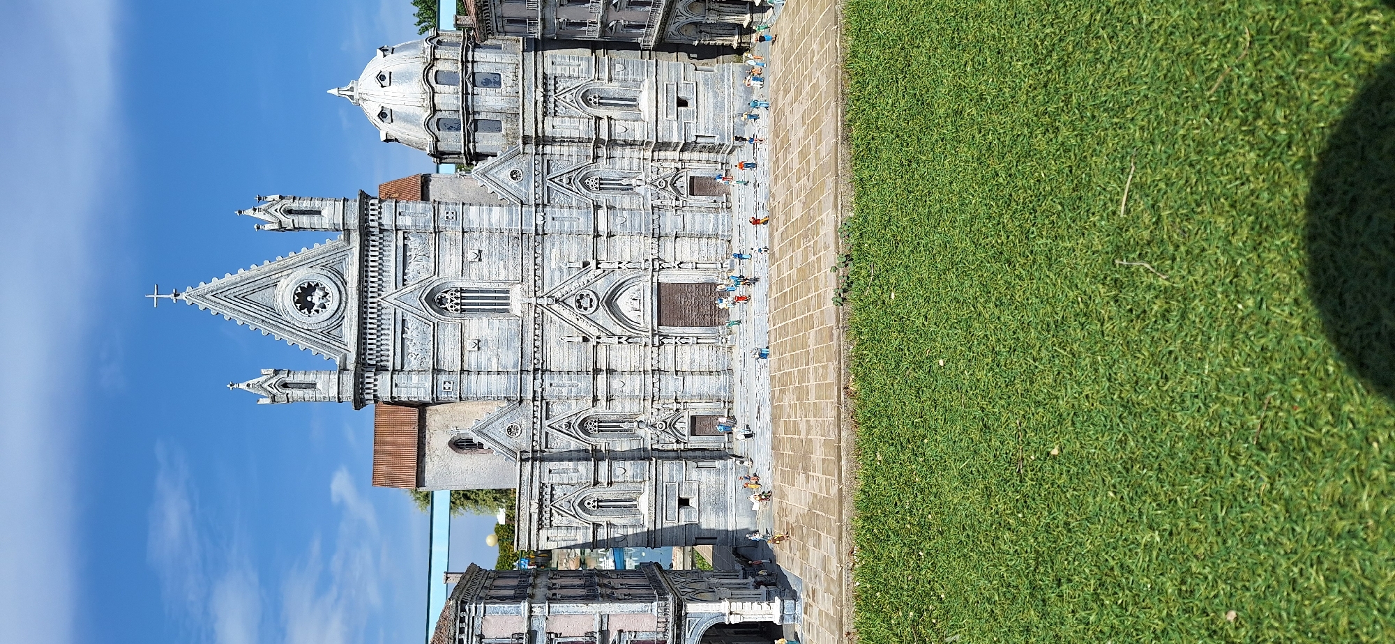 This image shows a miniature replica of the Duomo di Rimini, also known as the Malatesta Temple, located in Rimini, Italy. The model is situated outdoors with a bright blue sky and scattered clouds. In the foreground, a meticulously crafted green lawn is visible, with a few small twigs and shadows cast upon it. Behind the lawn, a tiled plaza represents the area in front of the cathedral. The miniature cathedral itself is the central focus, with its detailed facade, Gothic architecture, prominent cross, and a dome. Several tiny figures, representing people, are scattered across the plaza and the steps leading to the cathedral, giving a sense of scale to the model. The surrounding buildings in the miniature city are also depicted with architectural detail. The overall impression is a sunny day at an outdoor attraction showcasing historical architecture in miniature form.