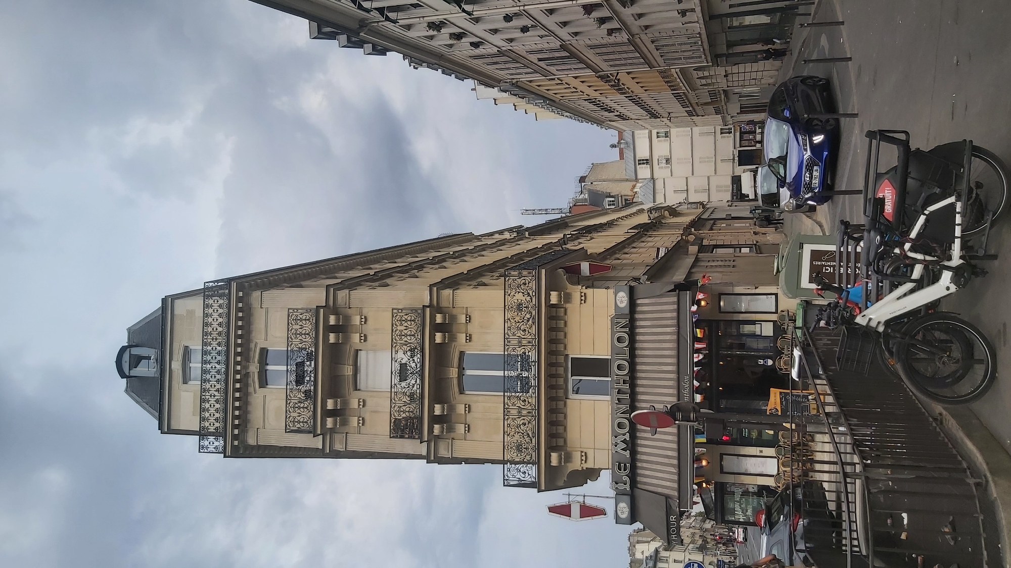 The image depicts a street scene in Paris, France, on a cloudy day. The foreground features two white bicycles parked on a sidewalk. To the left is a building with a distinctive angled facade, housing a cafe or restaurant named "LE MONTHOLON." The establishment has outdoor seating with chairs and tables. Various flags are strung across its awning. A black car is parked to the left of the cafe.
The midground shows a street with a blue Hyundai Ioniq car driving away from the camera. Several pedestrians are visible walking on the sidewalk and street. The background consists of Parisian architecture, with tall, narrow buildings lining both sides of the street. These buildings have multiple stories, French-style windows with shutters, and decorative balconies. A tall antenna or communication tower is visible in the distance. The sky is overcast with dense, grey clouds.