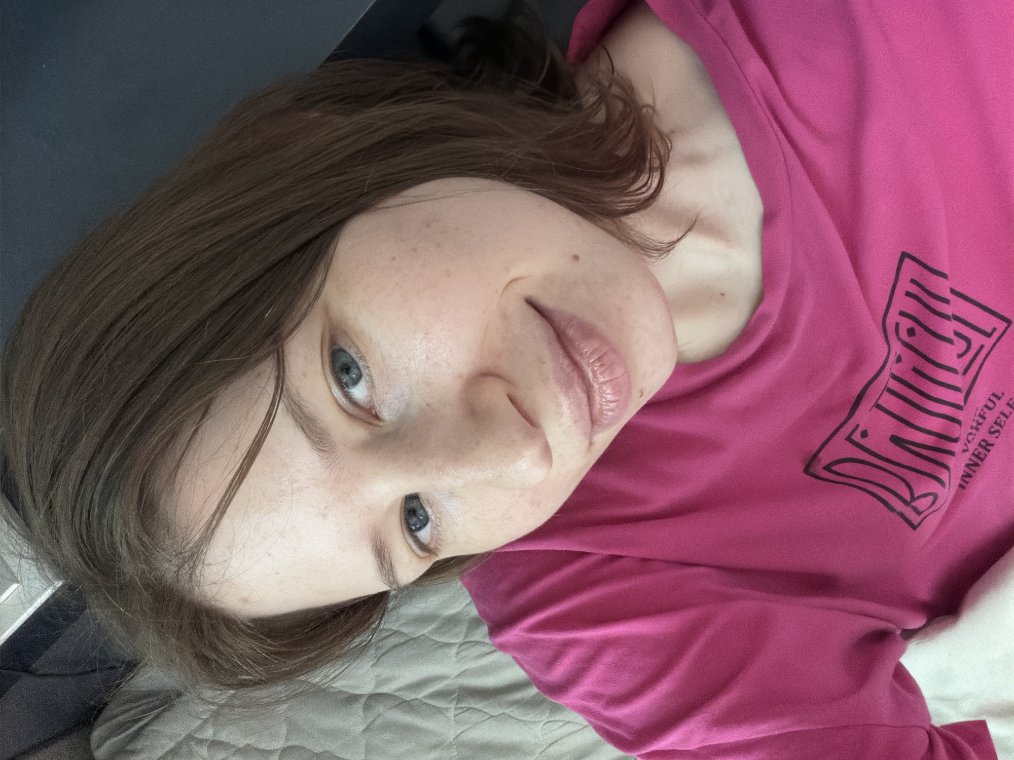 A young woman with shoulder-length brown hair and blue eyes, wearing a pink t-shirt with a black graphic print, is lying on a bed. She is looking up and to the right, with a slight smile on her face. The background shows a quilted white bedspread and a dark gray surface, possibly furniture. The lighting suggests it is daytime. The image does not contain information about the location in Dongducheon-si, South Korea.