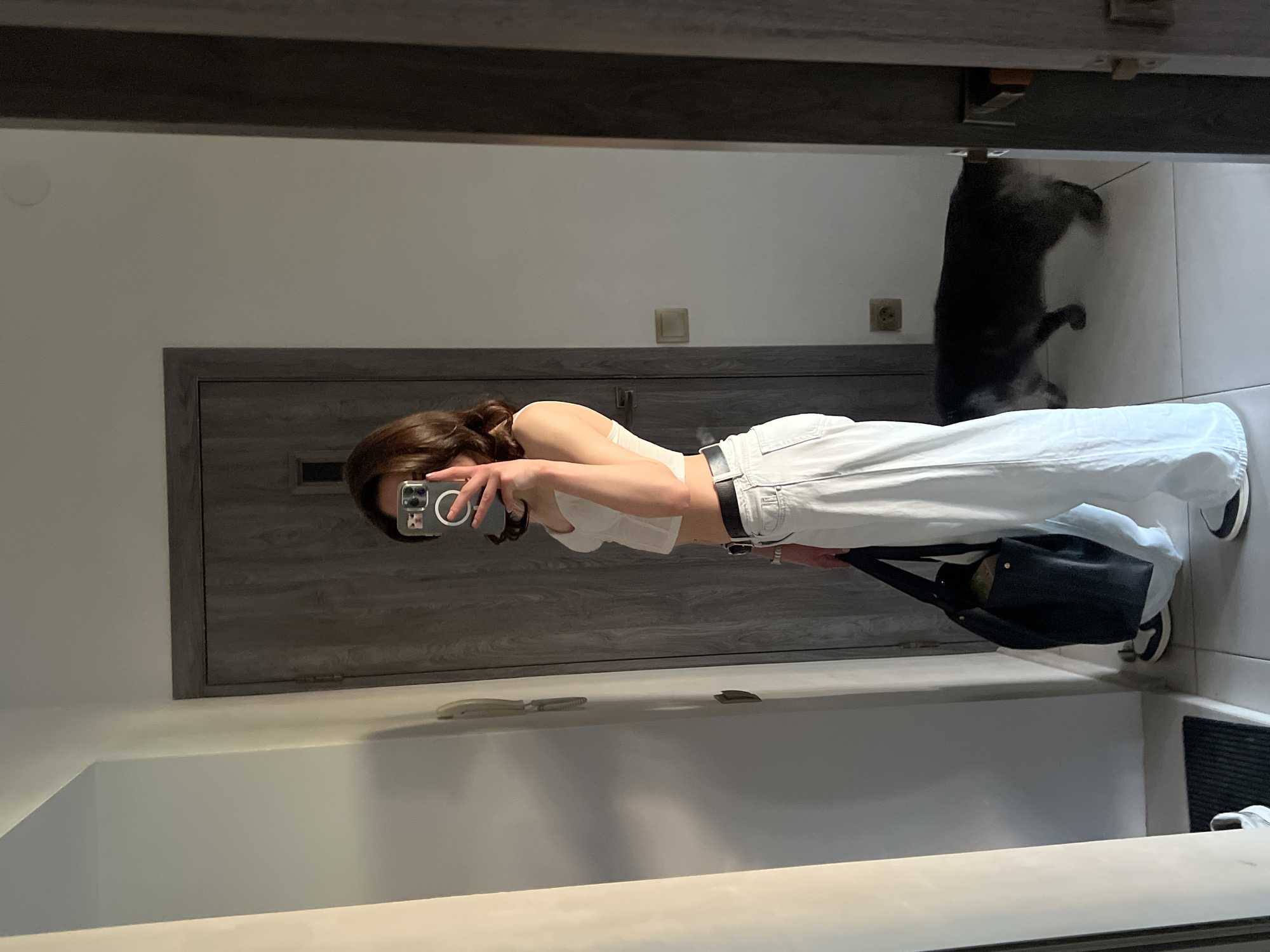 A young woman with dark, shoulder-length hair stands in a hallway, taking a mirror selfie with her phone. She is wearing a white cropped tank top, loose-fitting light blue jeans, a black belt, and black and white sneakers. She is holding a large black tote bag in her left hand. To the right of the woman, a grey cat walks away from the camera. The hallway features a grey wooden door, white walls, and tiled flooring. There is a door chime on the left wall. The scene appears to be in an apartment in Ternopil, Ukraine.