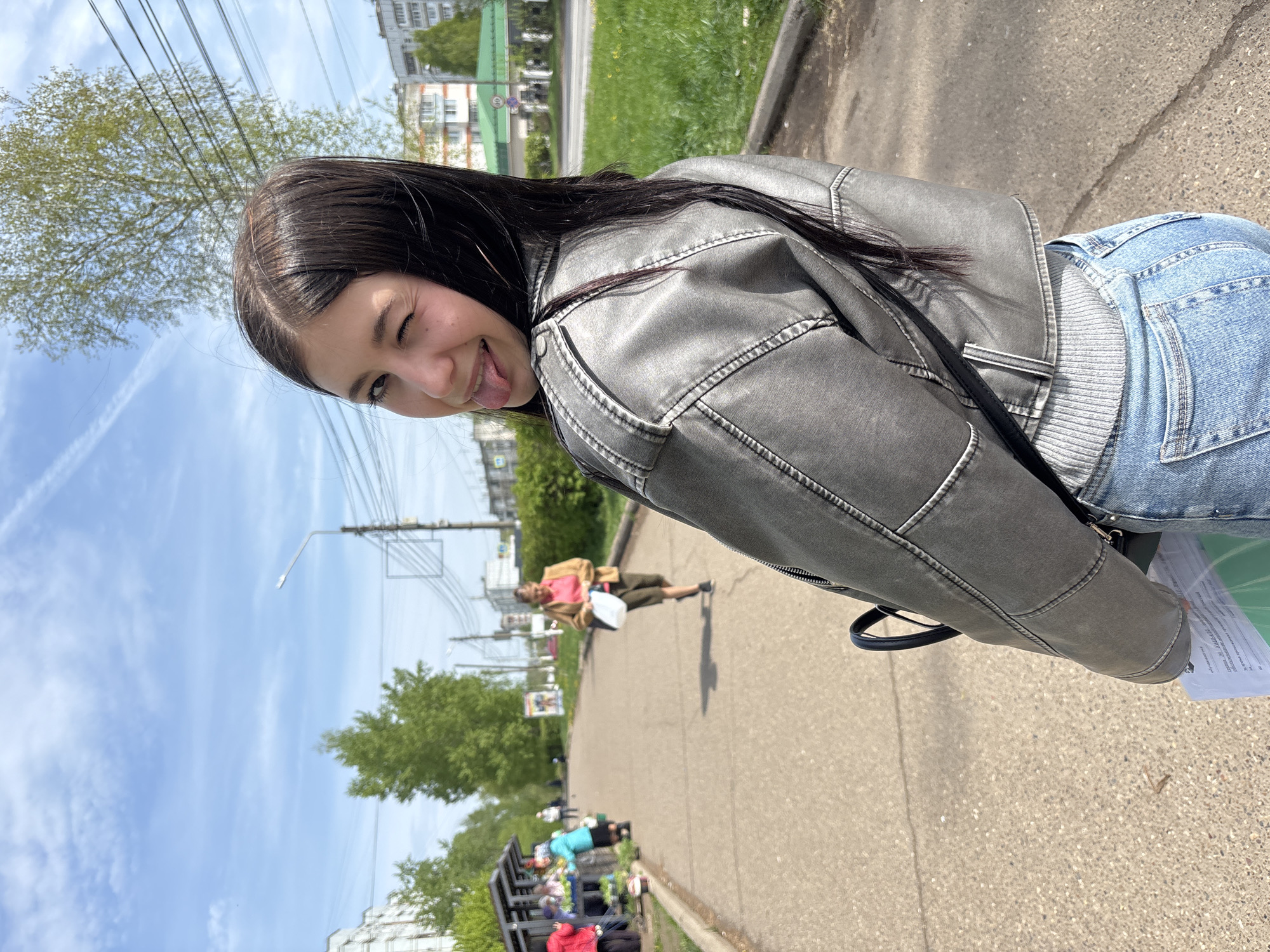 A young woman stands on a sidewalk in Chaikovsky, Russia, wearing a grey leather jacket and blue jeans, her tongue sticking out and one eye winking. In the background, a street with buildings and trees is visible. A man walks further down the sidewalk, and a bench with people is seen in the distance. The sky is blue with some clouds, and utility poles and wires are overhead.