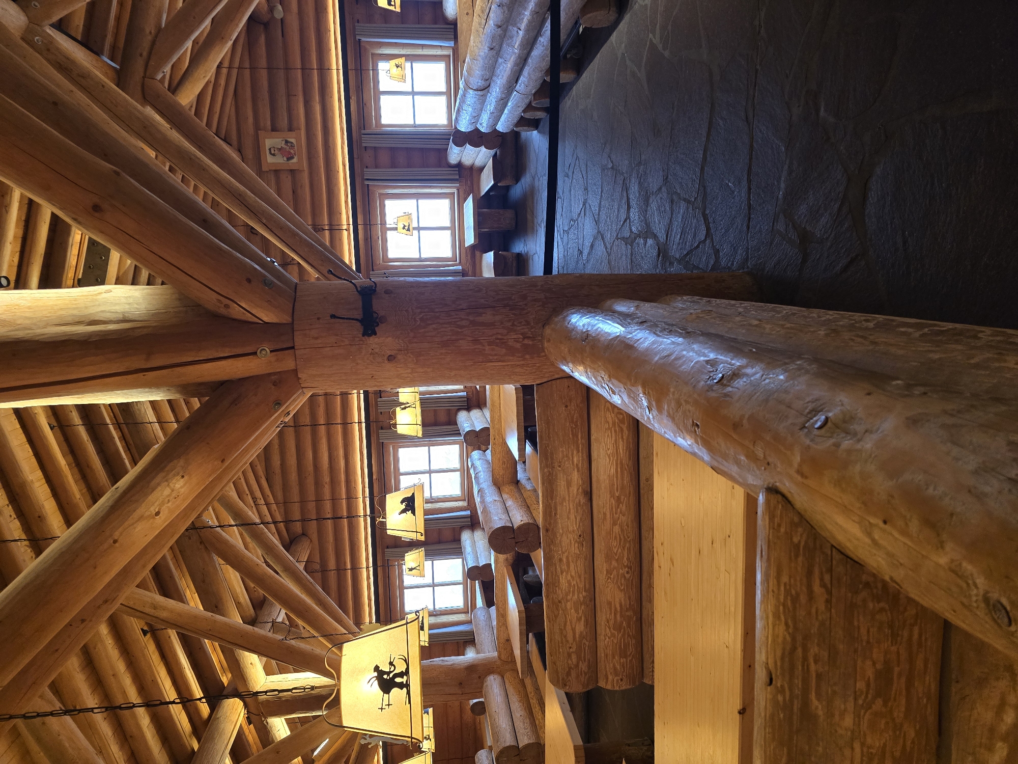 The image depicts the interior of a rustic restaurant constructed primarily from logs, reminiscent of a mountain lodge or a traditional Bavarian establishment, likely located in Garmisch-Partenkirchen, Germany. The ceiling features an exposed log beam structure with a pitched roof, and several wooden chandeliers with silhouette lamps hang from chains. The walls and seating areas are made from rounded logs, forming booths and benches with wooden tabletops. Large windows with white curtains provide natural light, revealing a glimpse of an outdoor setting. The floor is paved with irregular, dark stone tiles. In the background, on a wall, a small framed portrait of a man in historical attire is visible. The overall ambiance is cozy and natural, with the warm tones of the wood dominating the scene. There are no people or visible actions occurring in the image; it is a static view of the restaurant's interior.