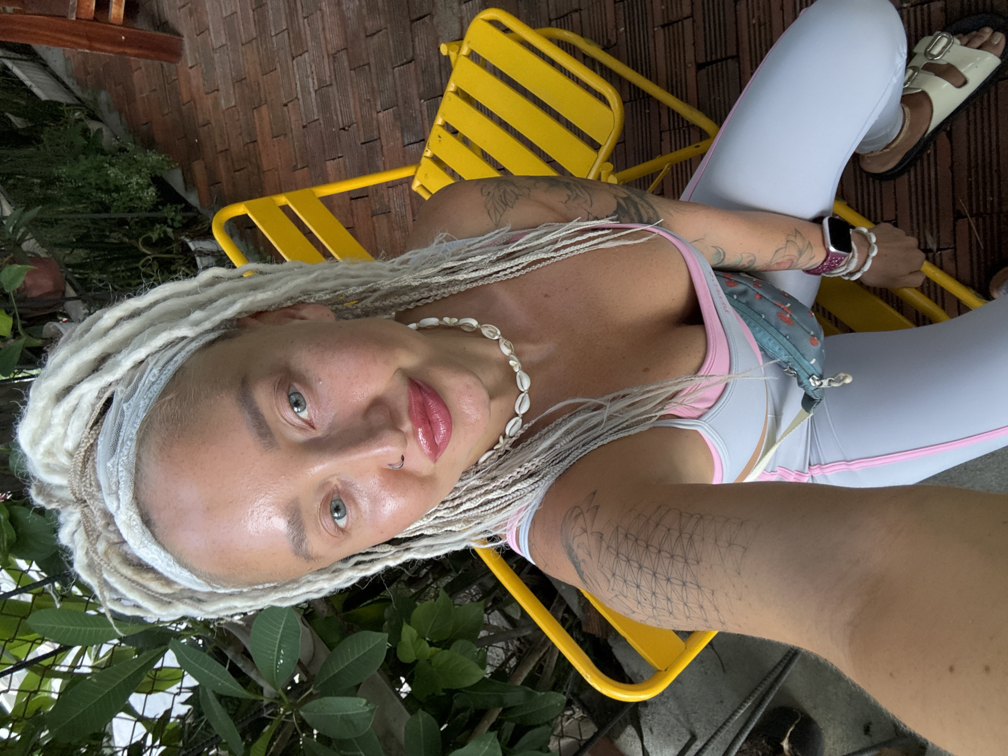 A woman with blonde dreadlocks and a shell necklace is seated on a yellow metal chair outdoors. She is wearing a light grey and pink sports bra and light grey leggings. She has tattoos on her arms and chest. A small blue pouch with a strawberry pattern is attached to her bra strap. She is looking at the camera with a slight smile. Her face is shiny, and her lips are bright pink. She is wearing a silver headband. Her right foot is visible, wearing a cream-colored sandal with two buckles. The background shows a tiled patio, greenery, and a hint of wooden structures. The scene appears to be outdoors during the daytime.