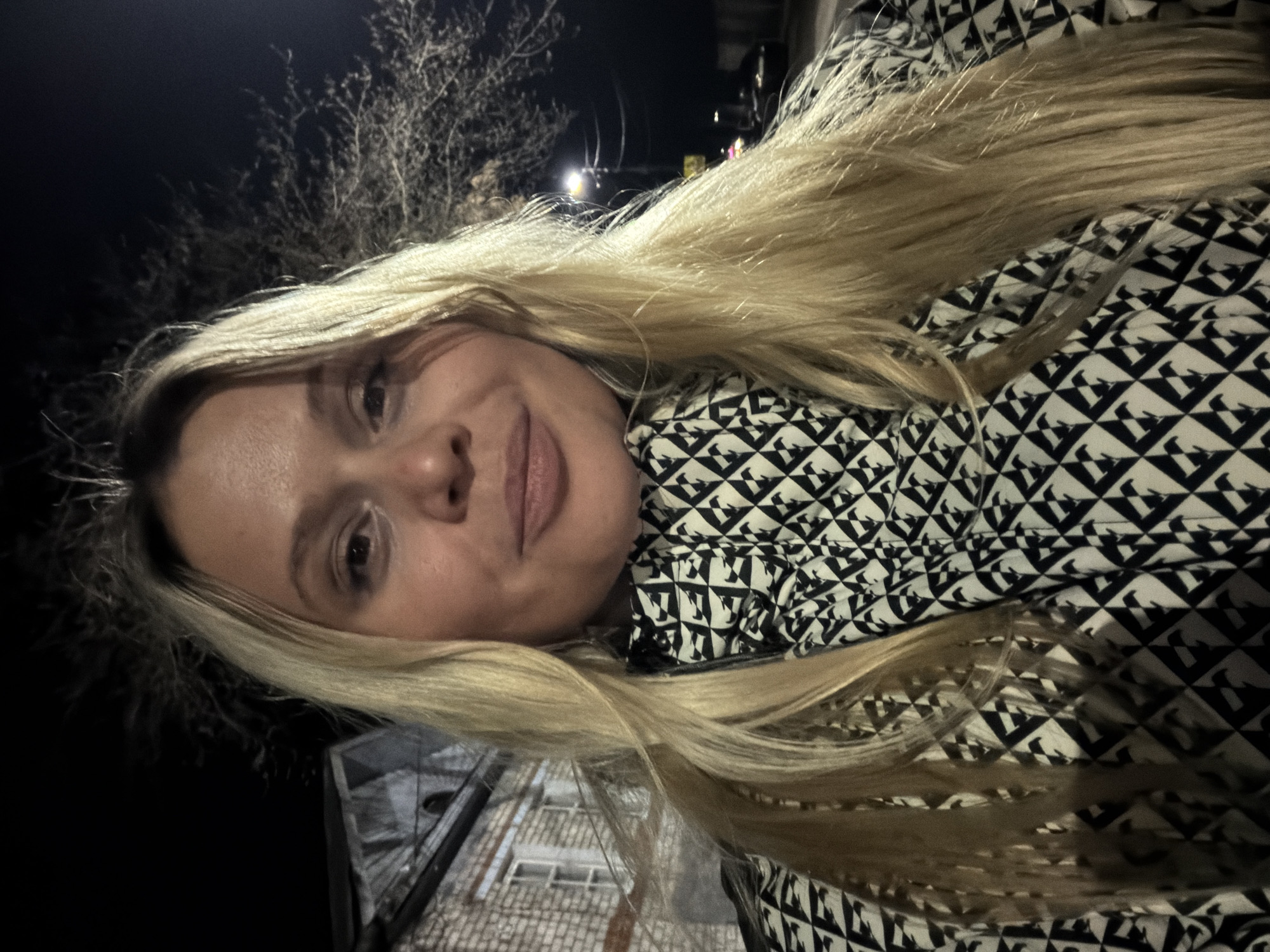 A selfie of a woman with long blonde hair is oriented horizontally and slightly skewed clockwise. She is wearing a black and white patterned jacket or coat. The background suggests an outdoor setting at night, with sparse trees and buildings. The location appears to be urban, possibly in the city of Sarapul, Russia, based on the prompt, although this cannot be definitively confirmed from the image alone.
