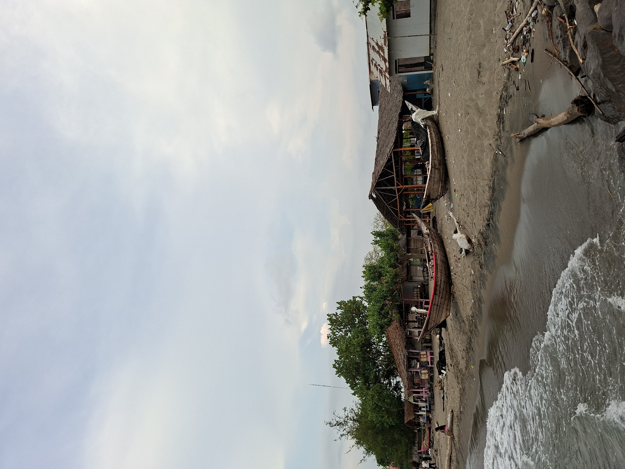 The image depicts a beach scene in Lhokseumawe, Indonesia. The sky is overcast with visible cloud formations. The shoreline has dark sand. The ocean water is gray with white foamy waves. Several wooden boats, some overturned, are beached along the shore. Behind the boats are buildings, some appearing to be open-air structures with thatched roofs, possibly restaurants or kiosks. One of the buildings appears to be made from corrugated metal. Debris and driftwood are scattered on the sand. A few people are visible on the beach further away from the camera.