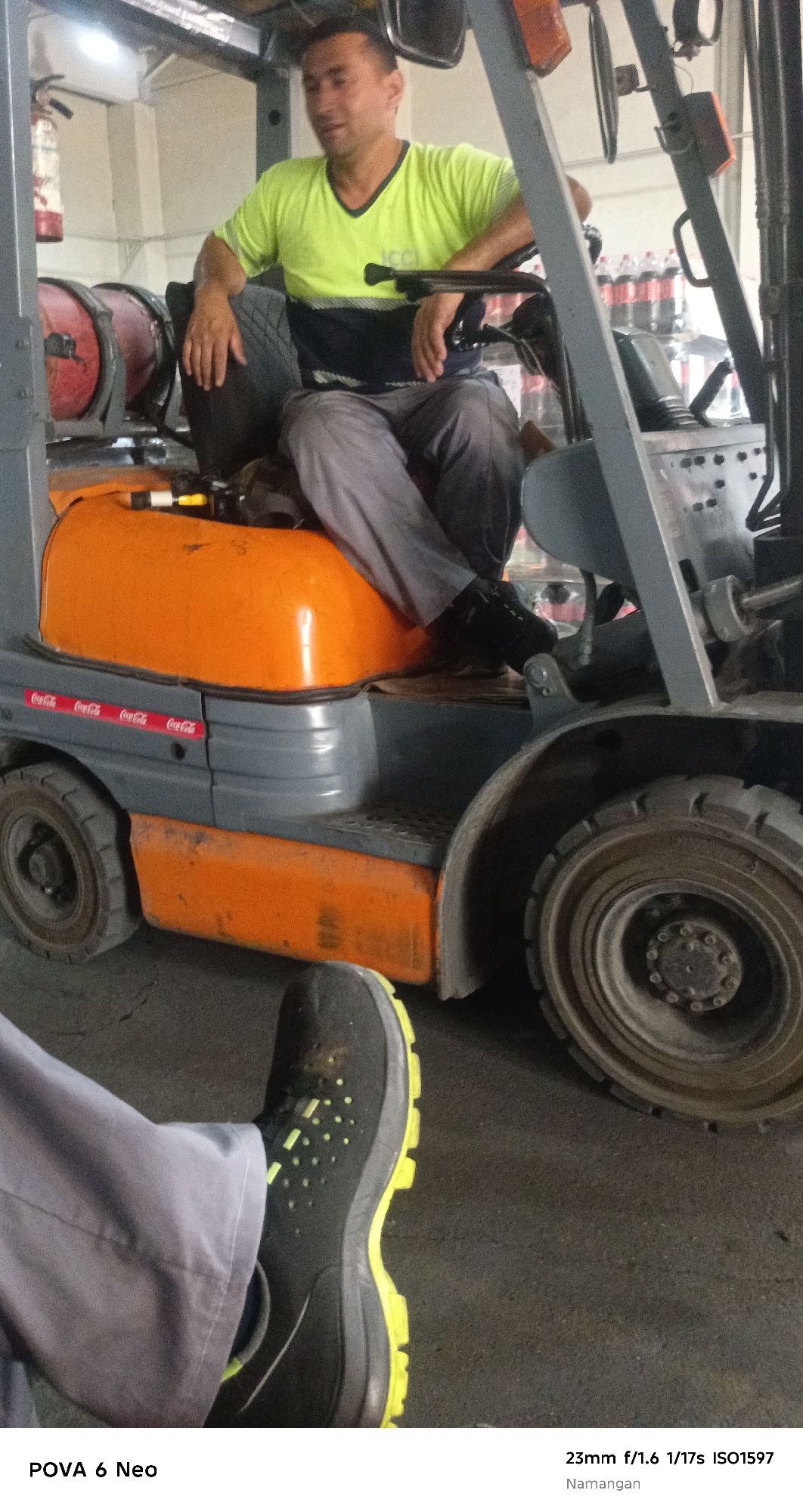 In Namangan City, Uzbekistan, a man wearing a lime green t-shirt with a dark blue band and grey pants is seated in the driver's seat of a forklift. The forklift has an orange and grey body, with the Coca-Cola logo visible on its side. The man appears to be looking to his right. In the foreground, a black shoe with yellow accents is visible, suggesting another person is present but not fully in frame. The environment appears to be an indoor industrial setting, possibly a warehouse or factory, with equipment and possibly shelves in the background. A fire extinguisher is mounted on a wall in the background.
