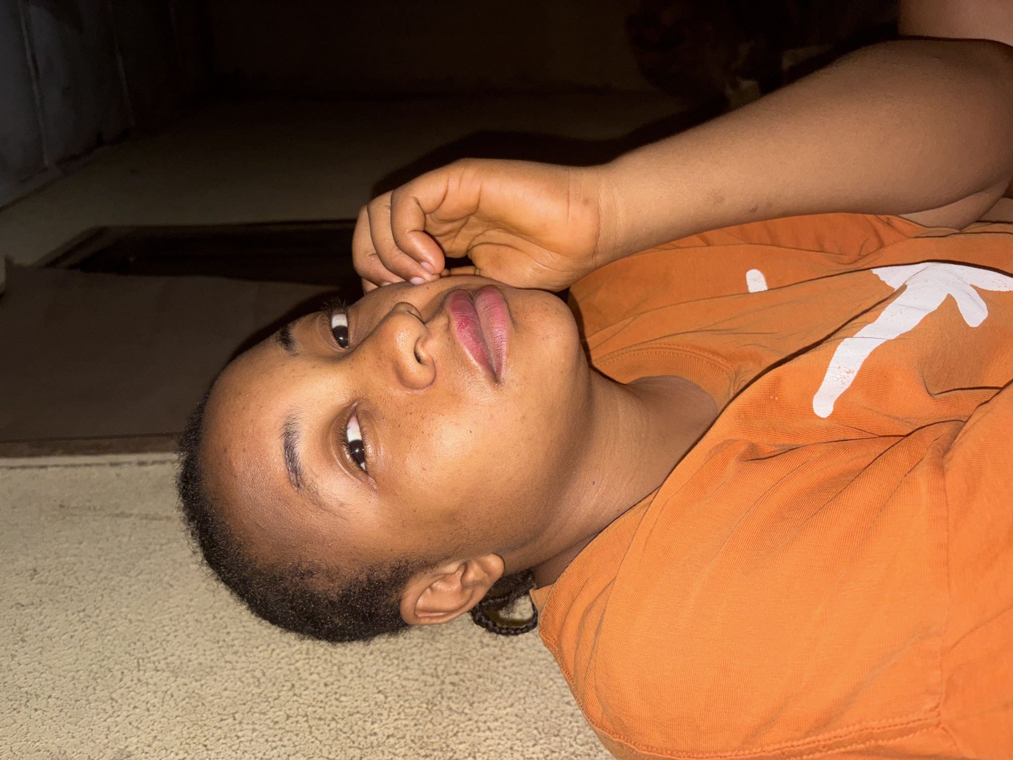 The image shows a young person with dark skin, likely female, lying on their side indoors. They are positioned with their head tilted towards the viewer's right, looking upwards with a calm expression. One hand rests near their chin and mouth. The individual has short, dark hair, dark eyes, and subtle freckles across their nose and cheeks. Their lips have a pinkish hue.
They are dressed in a bright orange long-sleeved top or hoodie featuring a partial white, abstract graphic on the front, which appears to be a stylized "V" or checkmark. The person is lying on a light-colored, possibly carpeted floor, which has a textured appearance. The background behind the subject is largely dark and indistinct, suggesting low-light conditions or a poorly lit room. The subject's face is well-illuminated by a direct light source. The scene depicts a relaxed pose in an indoor setting in Jos, Nigeria. No explicit text is visible, only the abstract white design on the shirt.