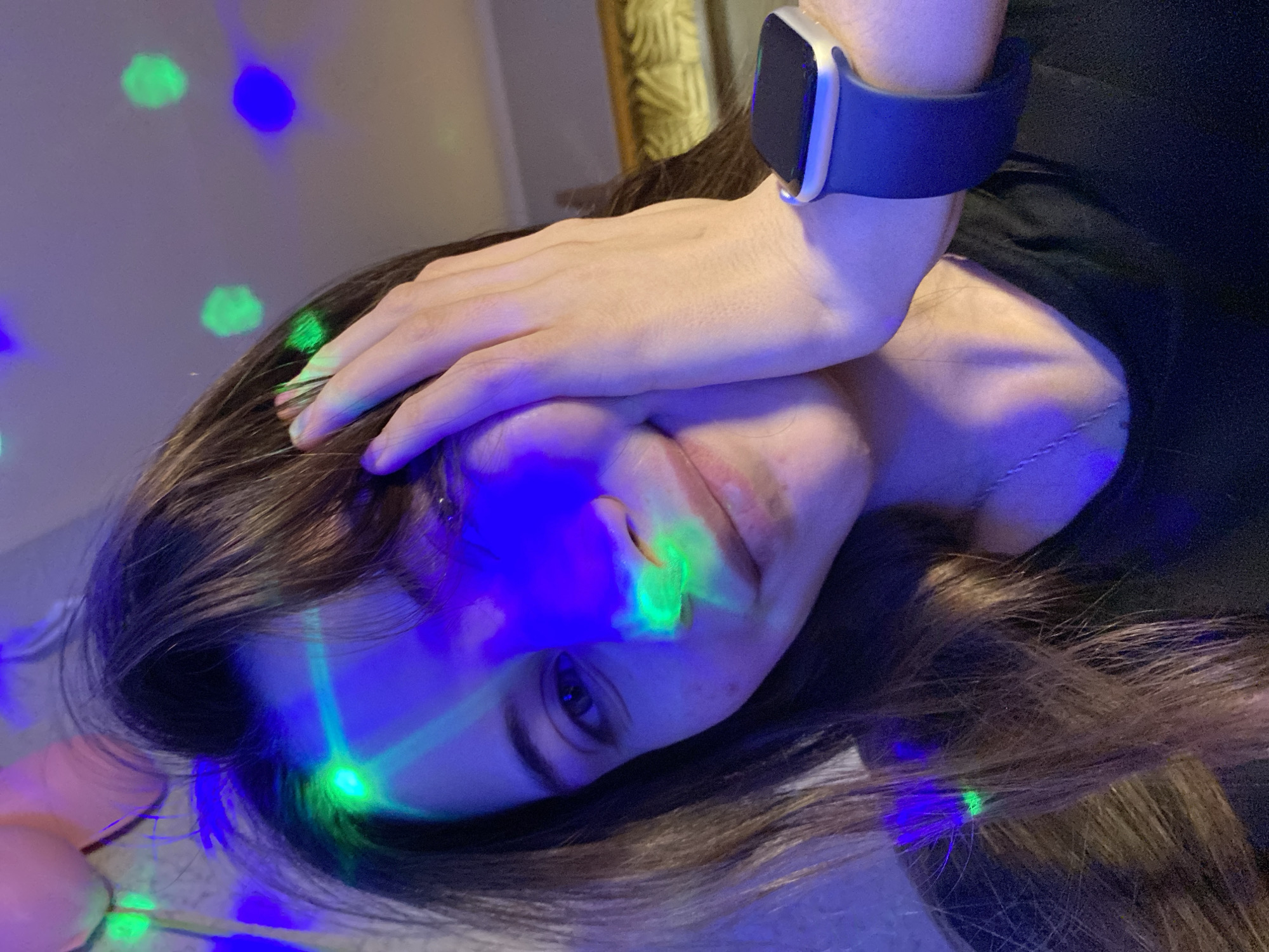 A woman is shown lying on her side, with her hand resting on her forehead. She is wearing a dark-colored top and a smartwatch with a blue band on her left wrist. The room is dimly lit with green and blue patterned lights. The location is unknown, but the prompt mentions the city of Votkinsk, Russia.