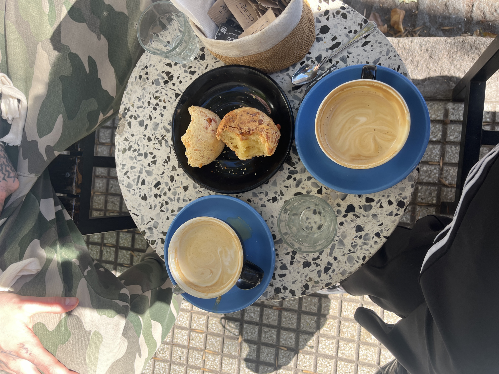 The image depicts a table at a cafe in Olivos, Argentina. The table, which has a light gray speckled surface, holds a black plate with two partially eaten cheese breads, two blue cups of coffee, one empty glass, one glass with remaining water, a small woven basket containing sugar packets, and one spoon. A person wearing camouflage pants is seated on the left, and another person wearing black Adidas pants with white stripes is seated on the right. The table is positioned outdoors on a tiled surface.