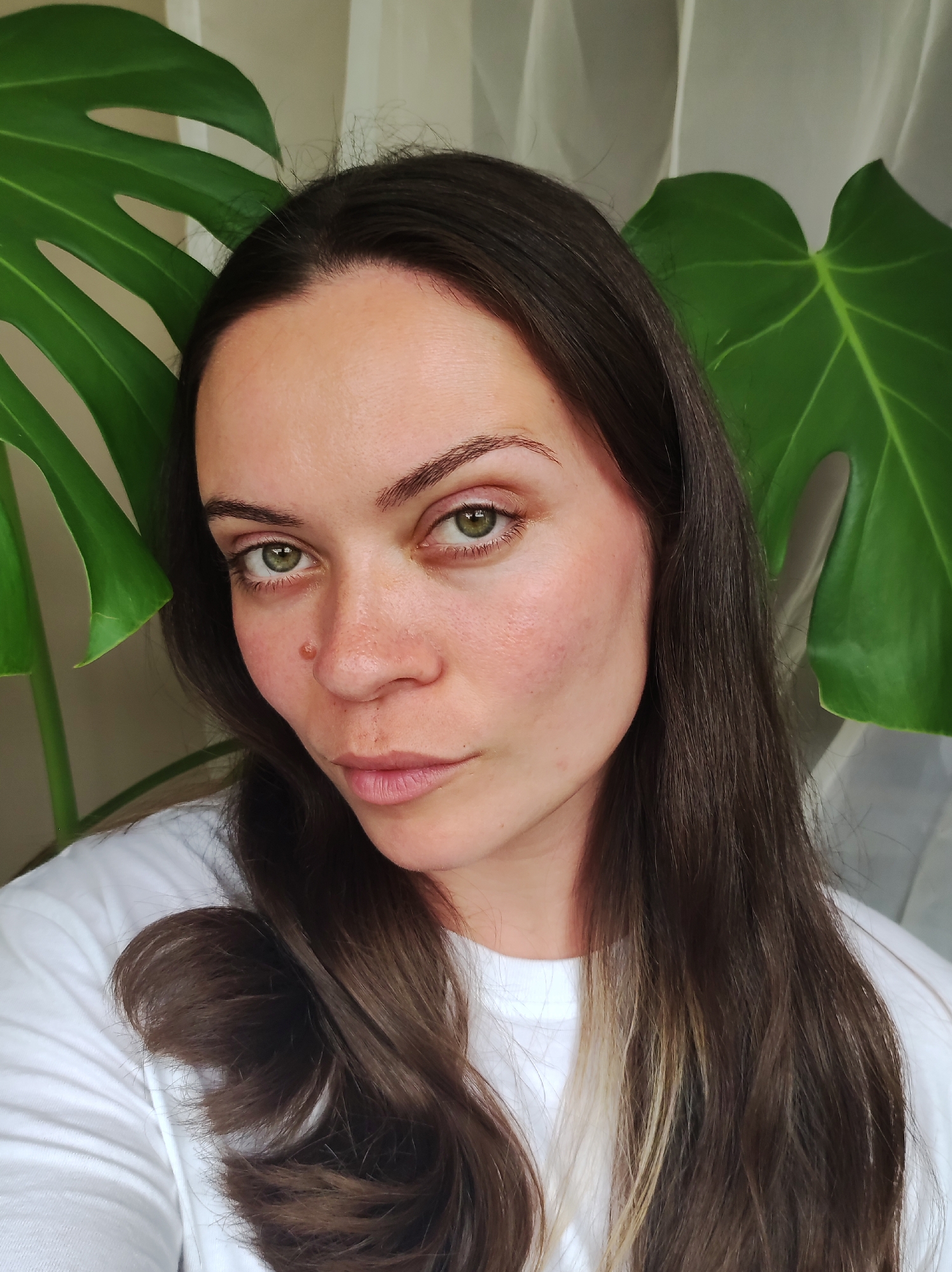 A woman with long, dark brown hair and green eyes looks directly at the camera. She is wearing a white t-shirt. Behind her, large green leaves of a Monstera plant are visible, along with a sheer white curtain. The scene appears to be indoors, possibly in a home or studio. The image does not provide any information about a specific location in the city of Chaikovsky, Russia.