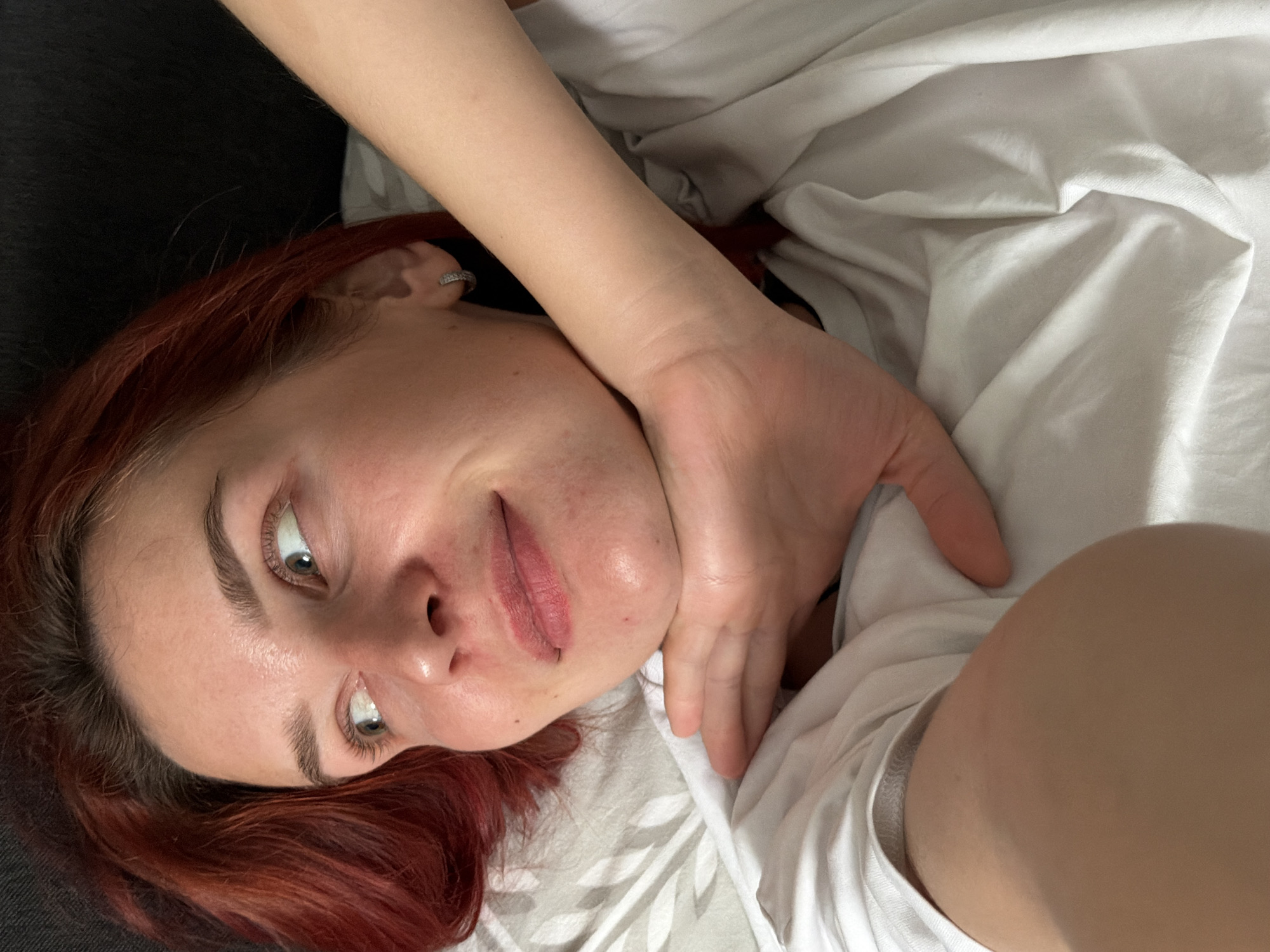 A woman with red hair lies on a patterned surface with a white shirt or sheet draped over her body. Her right hand is positioned near her neck. She appears to be indoors, likely in a domestic setting. The location in the city of Naberezhnye Chelny, Russia is not discernible from the image.
