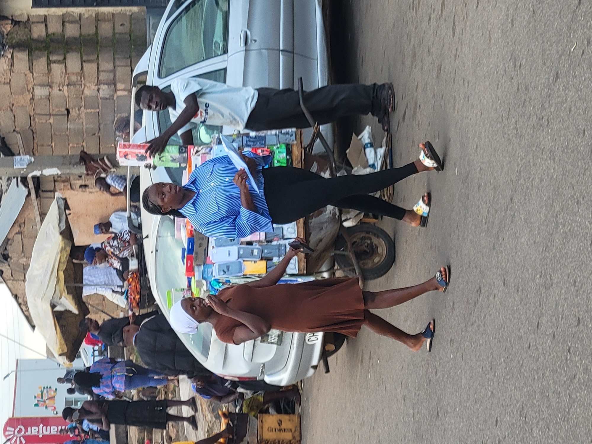In Barakin Lerawi, Nigeria, a street scene unfolds. A woman wearing a brown dress and white head covering walks with her hand to her mouth, appearing to be eating something. Beside her, a woman in a blue striped shirt and black leggings holds a clipboard. Behind them, a man in a light blue t-shirt holds up a boxed item. A silver car is parked nearby, and a cart laden with various goods, including water bottles and phone cases, is positioned behind the women. In the background, market stalls with an umbrella canopy and other pedestrians are visible. The environment suggests an outdoor market or street vendor setting.