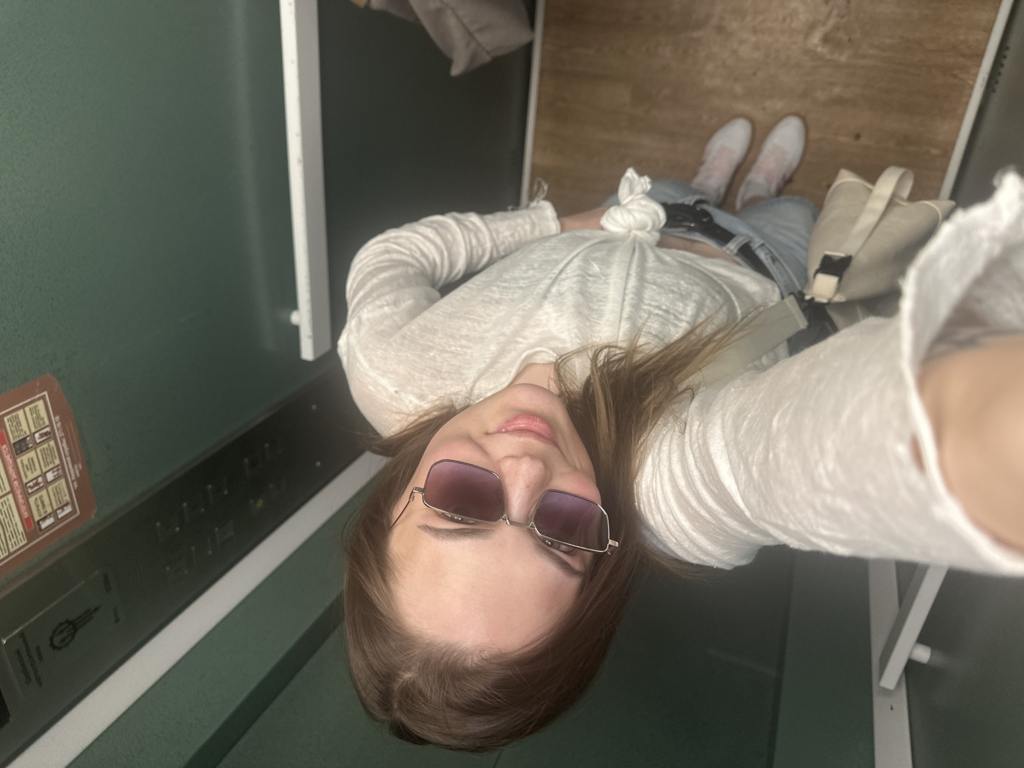 A selfie taken from above shows a woman in an elevator. The woman is wearing sunglasses, a white long-sleeved shirt tied at the waist, jeans, and white sneakers with a pink design. She is holding a light-colored purse or bag. The elevator has green walls and metal fixtures, including elevator buttons and a sign with information. The floor of the elevator is wood-like. It is not possible to determine if the location is in the city of Perm, Russia.